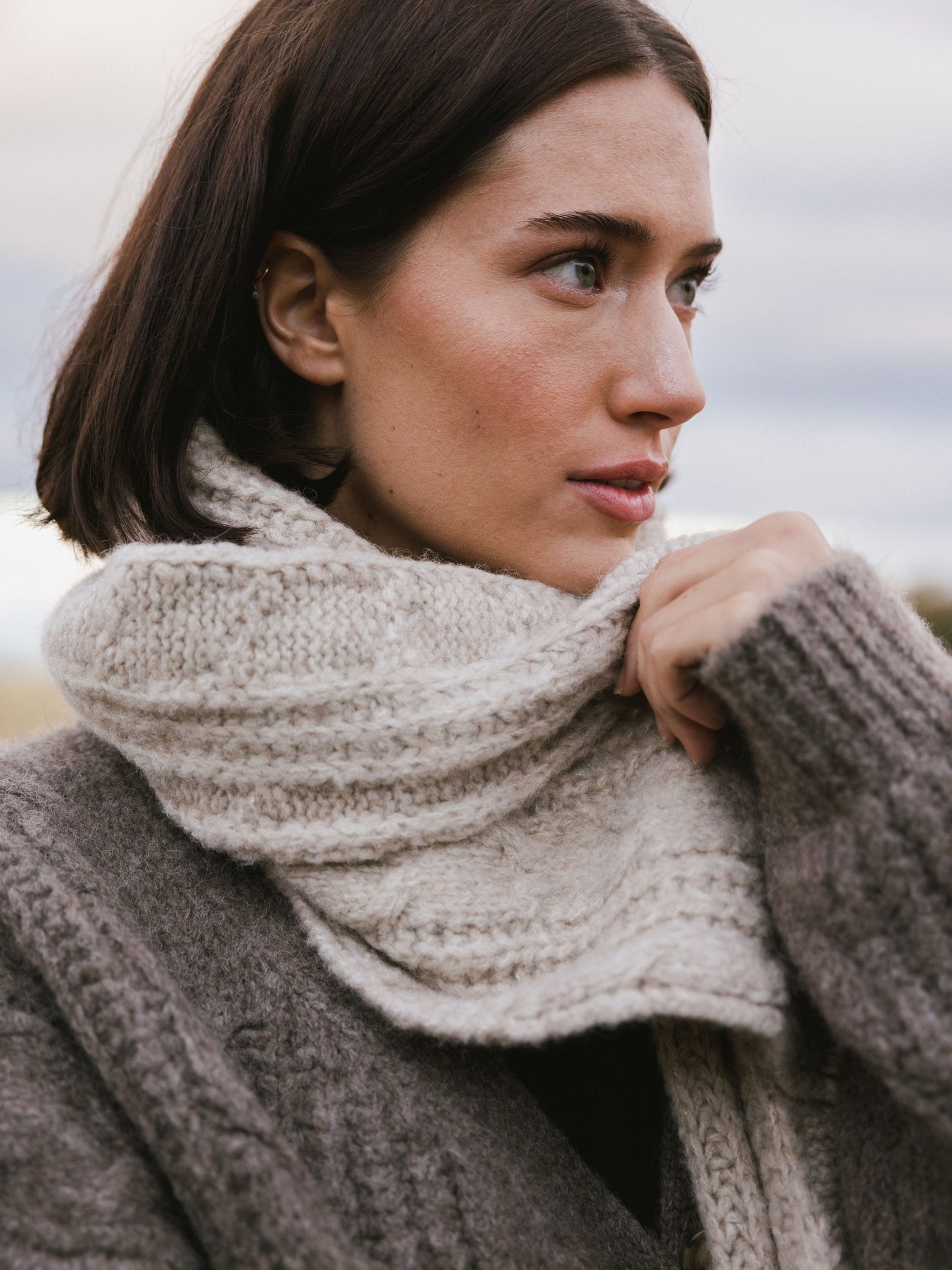 Woman wearing a brown knit cardigan, Cozy Earth Cable Knit Scarf in beige in a field.