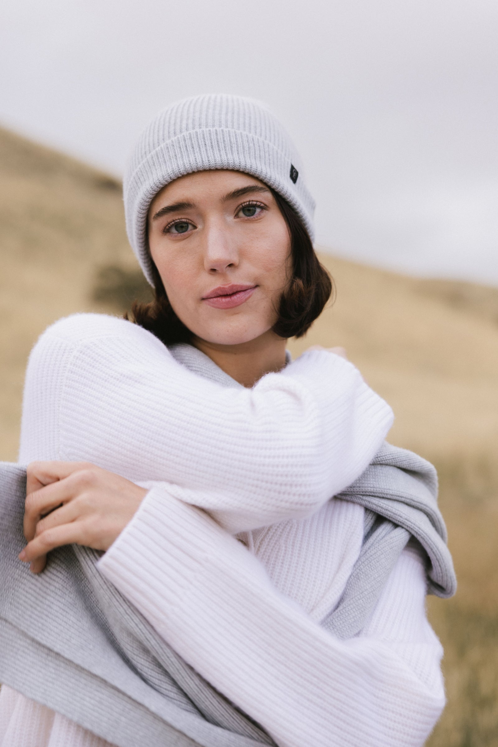 Woman wearing a light grey scarf, white sweater, and light-colored pants in a field