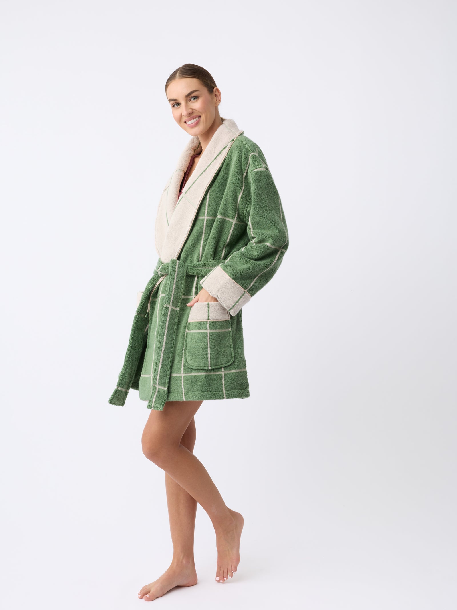 A smiling person stands barefoot in a Windowpane Resort Robe by Cozy Earth, featuring a green and white checkered pattern with cream-colored trim, posing against a plain white background.