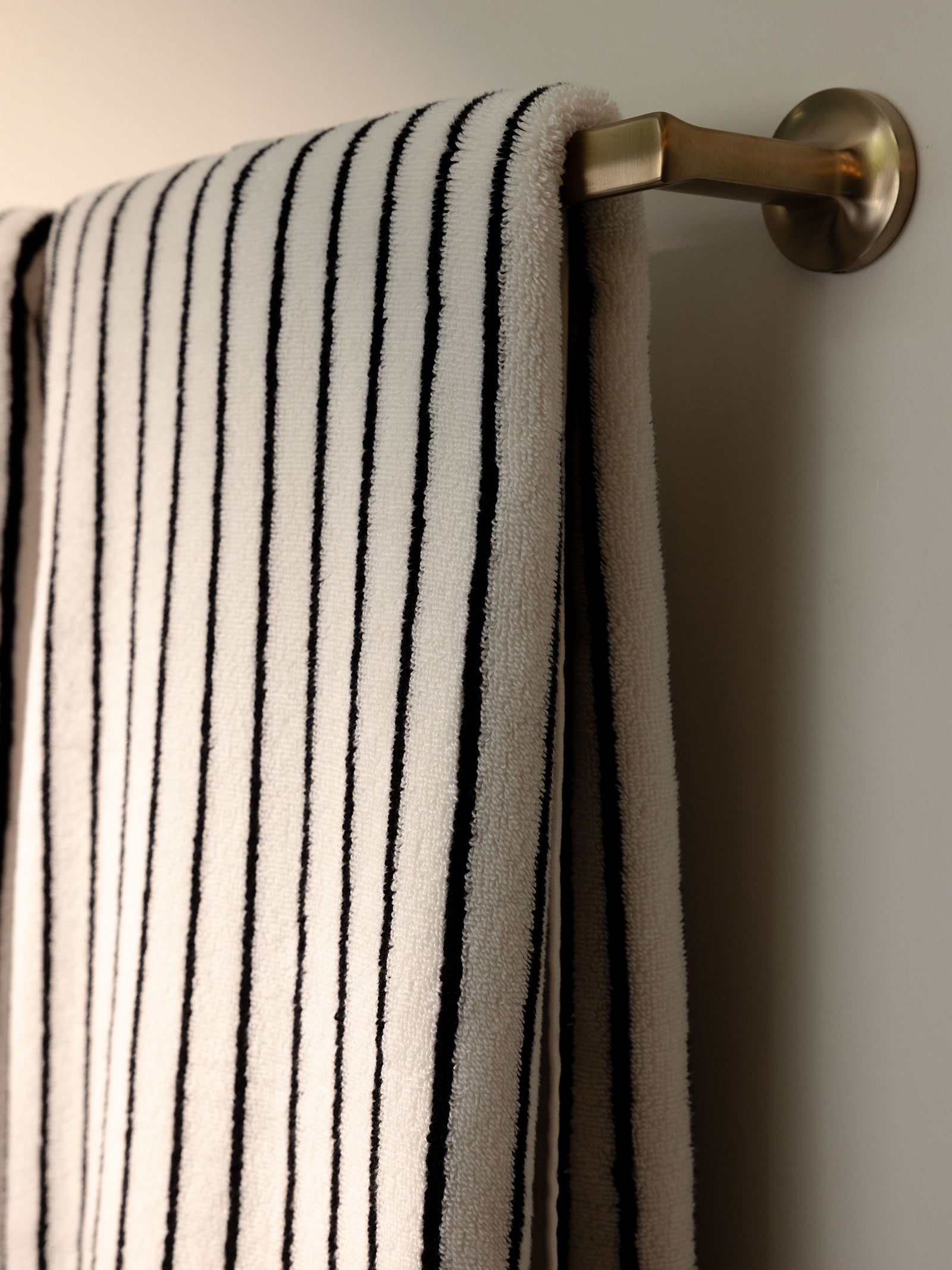 A Luxe Bath Towel from Cozy Earth, featuring elegant black vertical stripes on a pristine white fabric, hangs neatly on a brass towel rod mounted on a white wall. Soft lighting casts gentle shadows, accentuating the towel's plush texture.