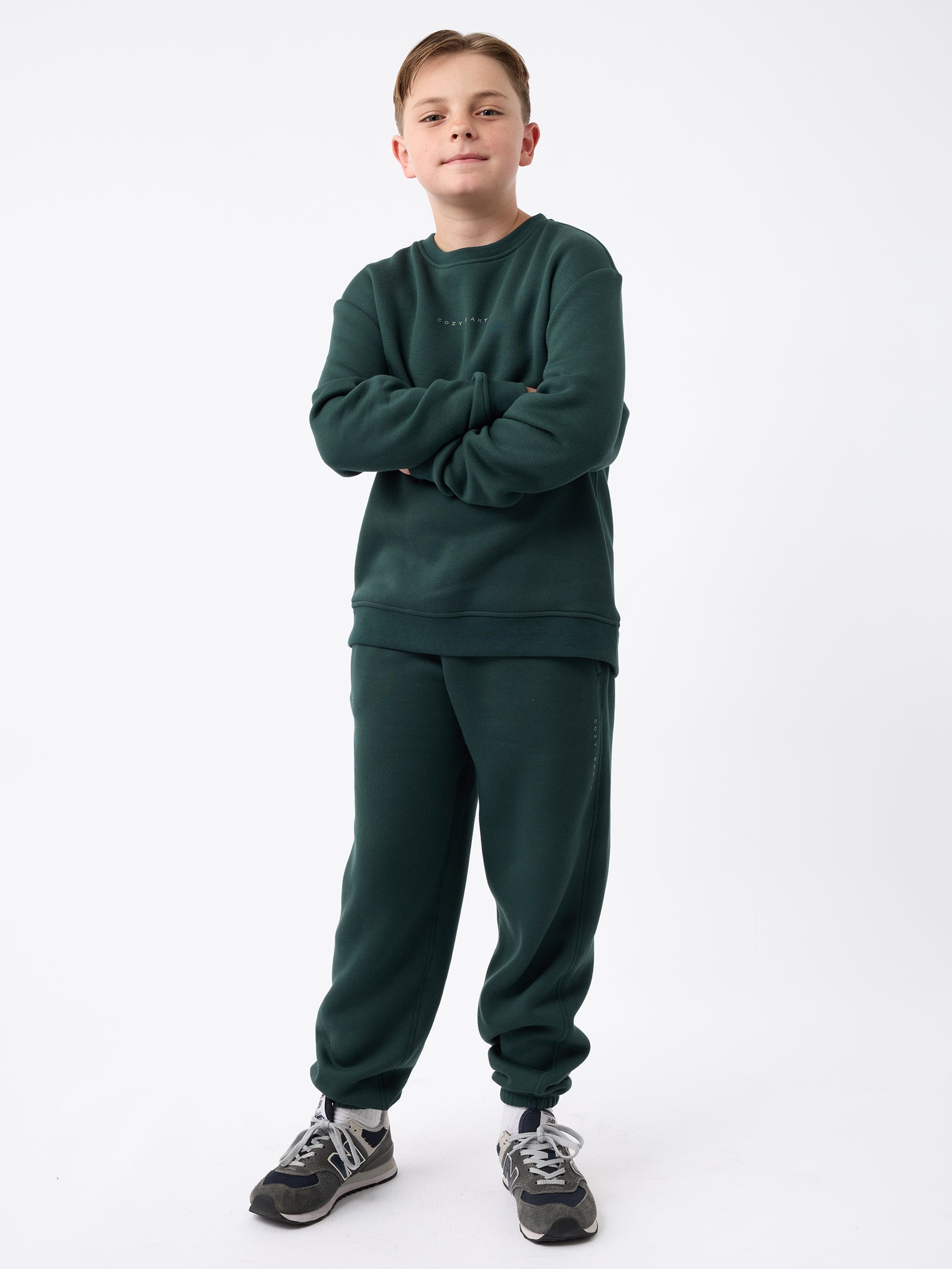 A young boy stands confidently with arms crossed, wearing Cozy Earth's Kid's CityScape Crewneck sweatshirt and matching sweatpants in dark green. With short brown hair and a slight smile, he complements his outfit with dark gray sneakers. The background is plain white.