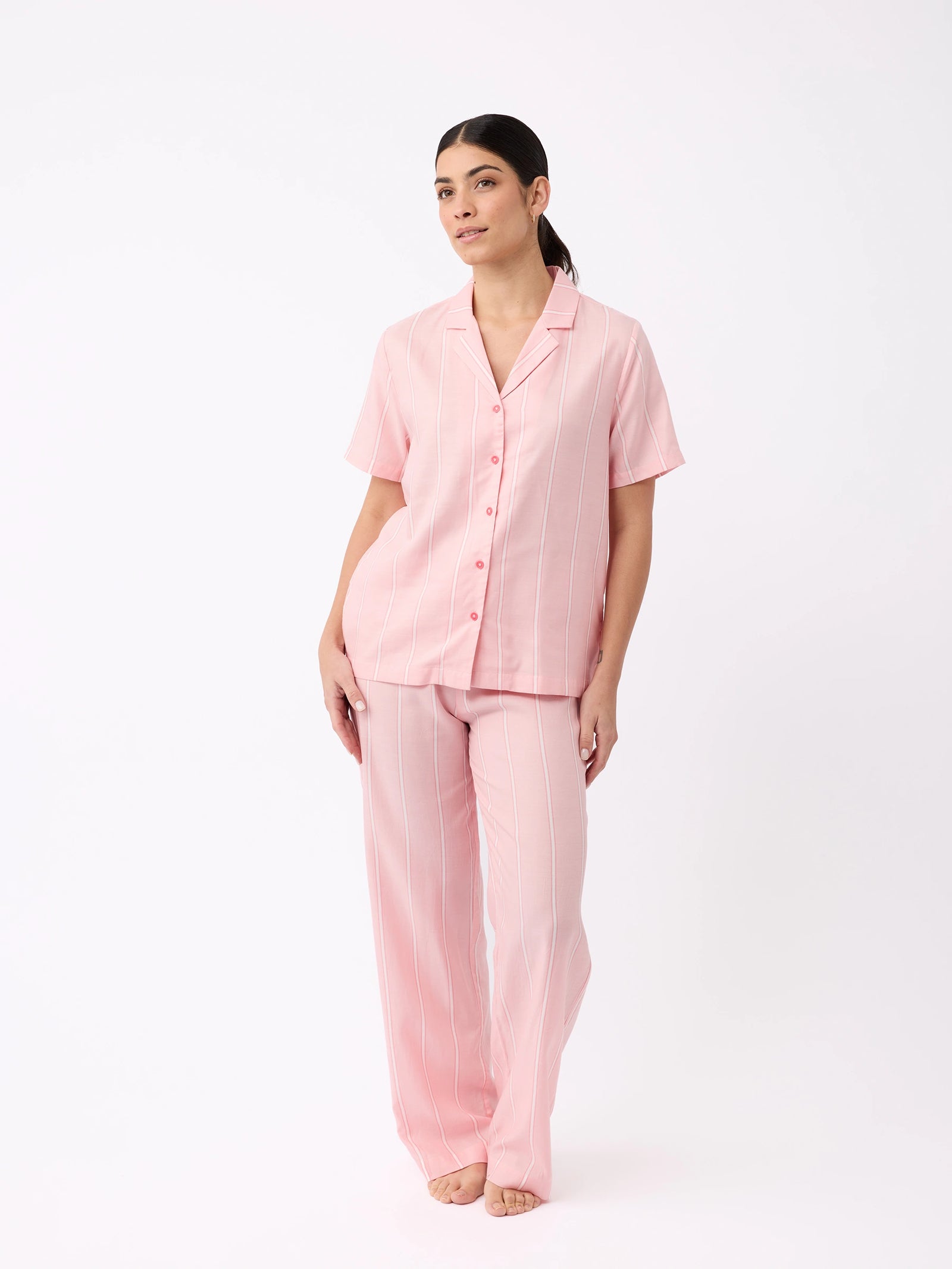 A person models a light pink, pinstriped pajama set from Cozy Earth. The short-sleeve top features buttons and the pants are relaxed fit, all posed against a white backdrop.