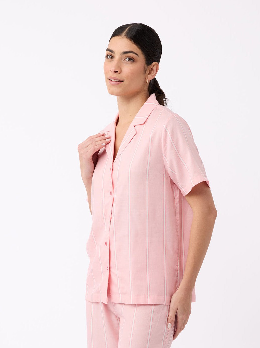 A woman with dark hair in a low ponytail wears the Cozy Earth Women's Soft Woven Short Sleeve Pajama Top in light pink with white stripes, standing against a plain white background and smiling softly.
