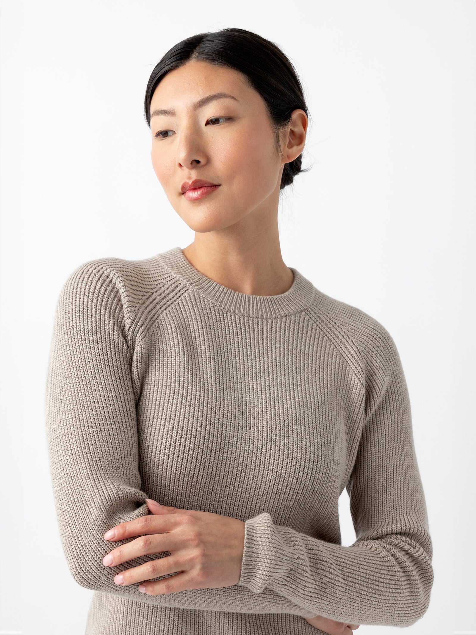 A person with dark hair pulled back is wearing a light gray, long-sleeved Women's Classic Crewneck from Cozy Earth. They are looking to the side with a neutral expression and have their arms crossed gently in front of them. The background is plain white.