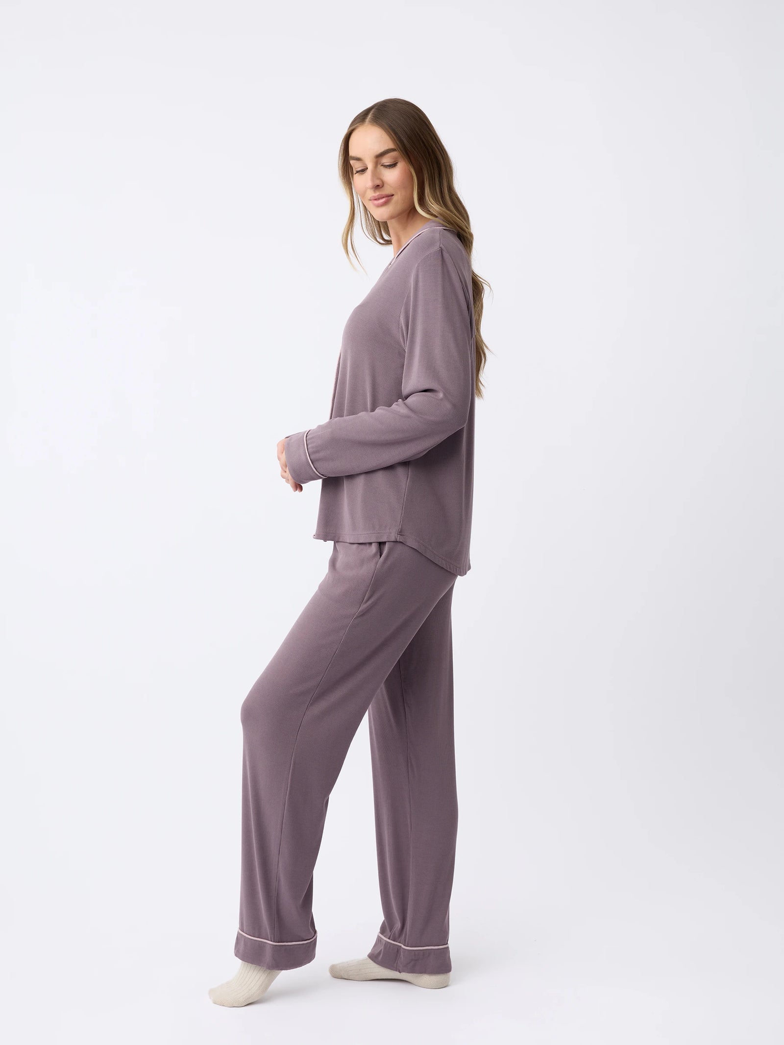 A woman with long hair stands sideways wearing the Rib-Knit Classic Long Sleeve Pajama Set by Cozy Earth. The matching gray outfit includes a long-sleeved top and pants. She looks down with a neutral expression against a plain white background.