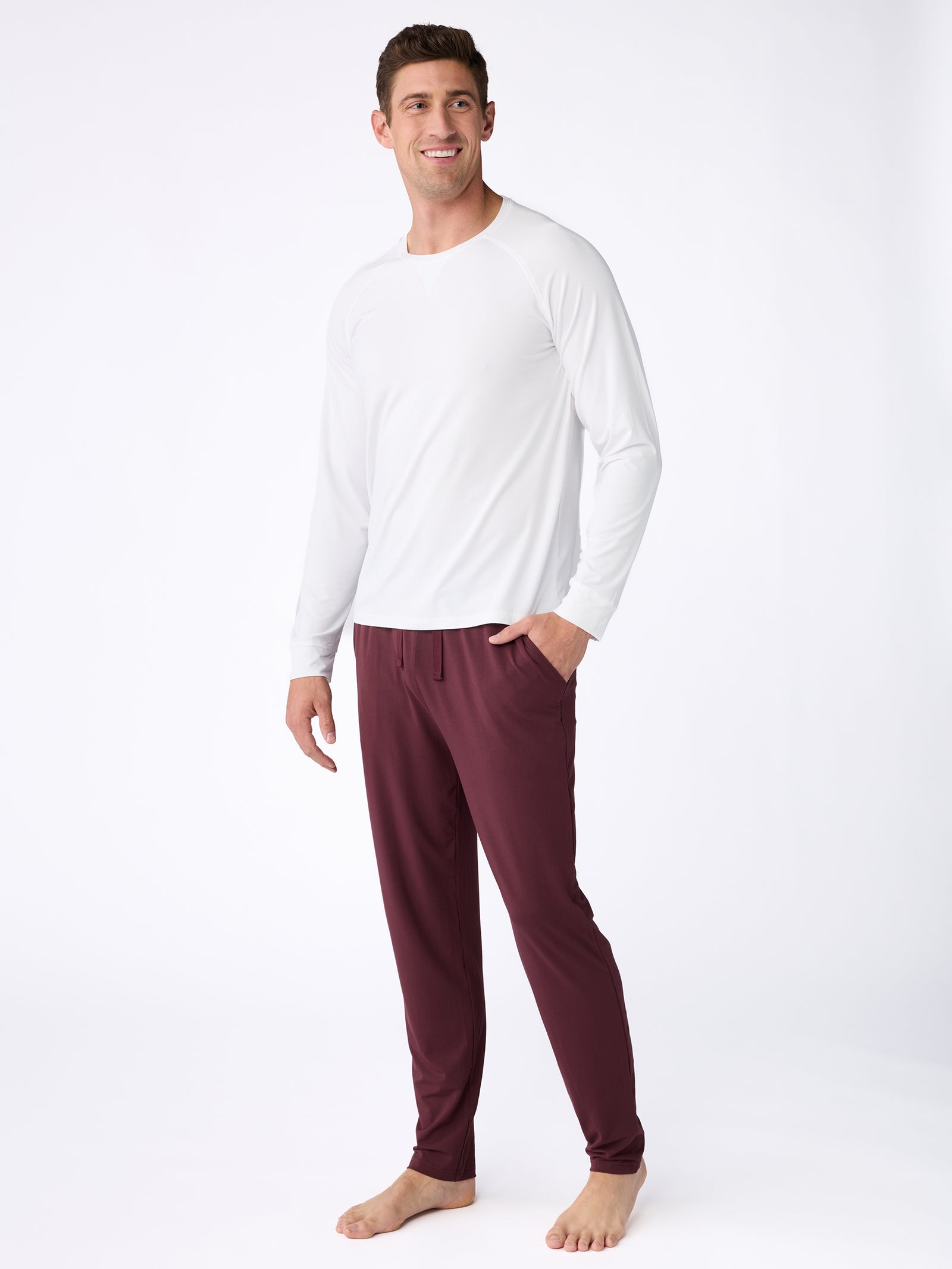 A person wearing the Cozy Earth Men's Stretch-Knit Bamboo Long Sleeve in white and maroon pajama pants stands barefoot against a plain white background, smiling with one hand in their pocket.