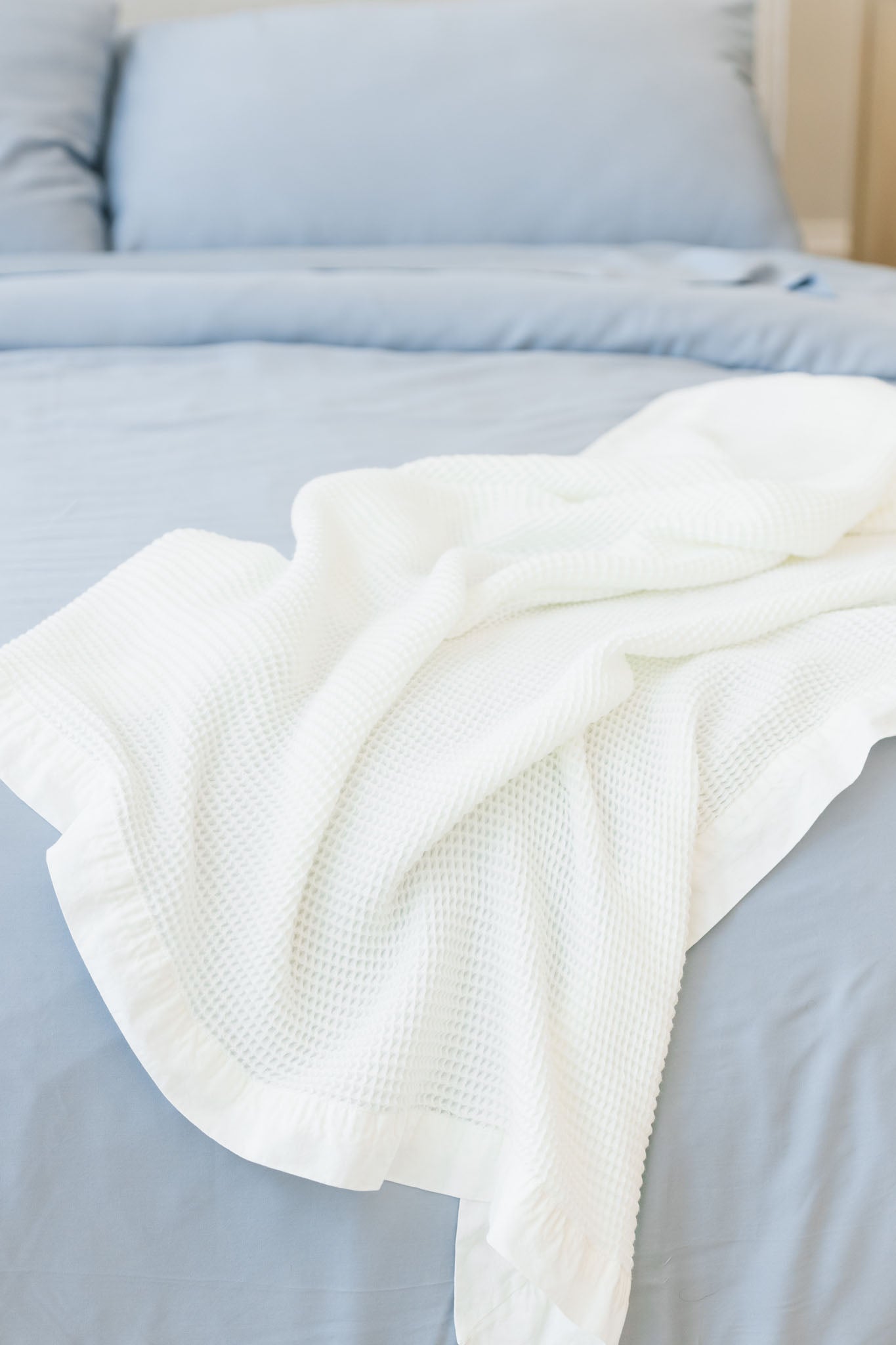 A Cozy Earth Waffle Blanket in white is casually draped over a light blue bedspread on a neatly made bed. The light blue pillows and bedspread provide a peaceful and serene backdrop to the textured blanket.