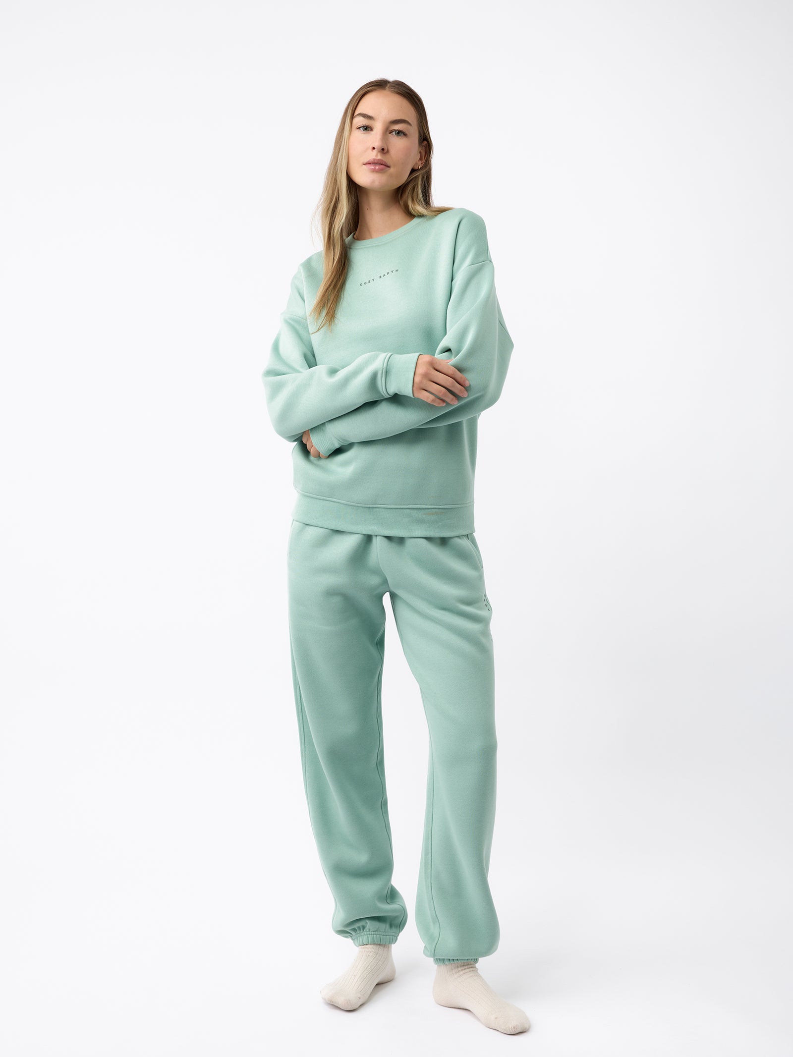 A woman wearing the Women's CityScape Crewneck by Cozy Earth stands gracefully in a light green sweatshirt and sweatpants. With long flowing hair and a direct gaze fixed on the camera, she embodies a casual and comfortable style, standing barefoot against a plain white backdrop.
