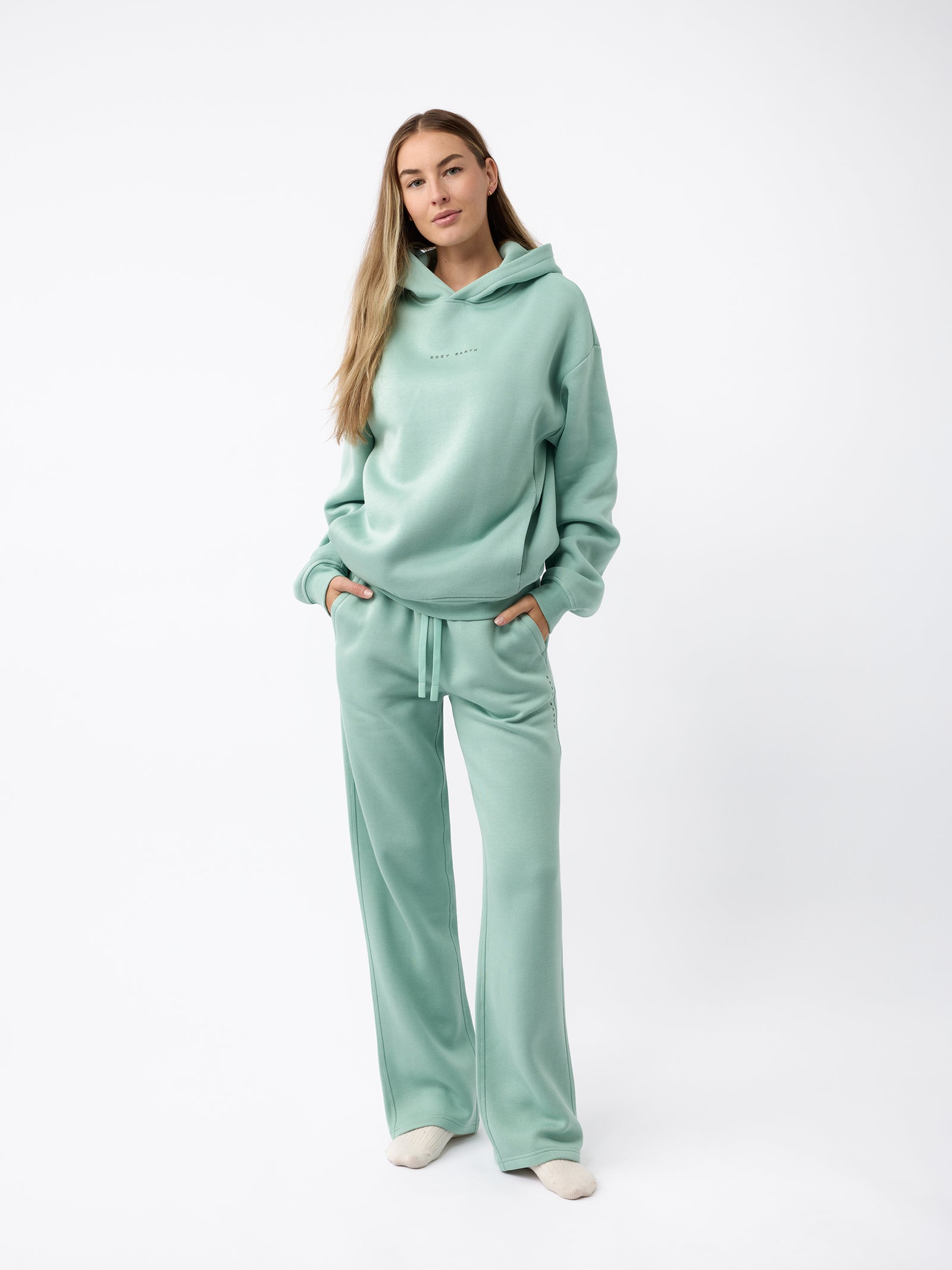 A person dressed in a mint green Women's CityScape Hoodie by Cozy Earth and matching joggers stands with hands in pockets against a plain white background. They have long, light brown hair and are sporting cream-colored shoes.
