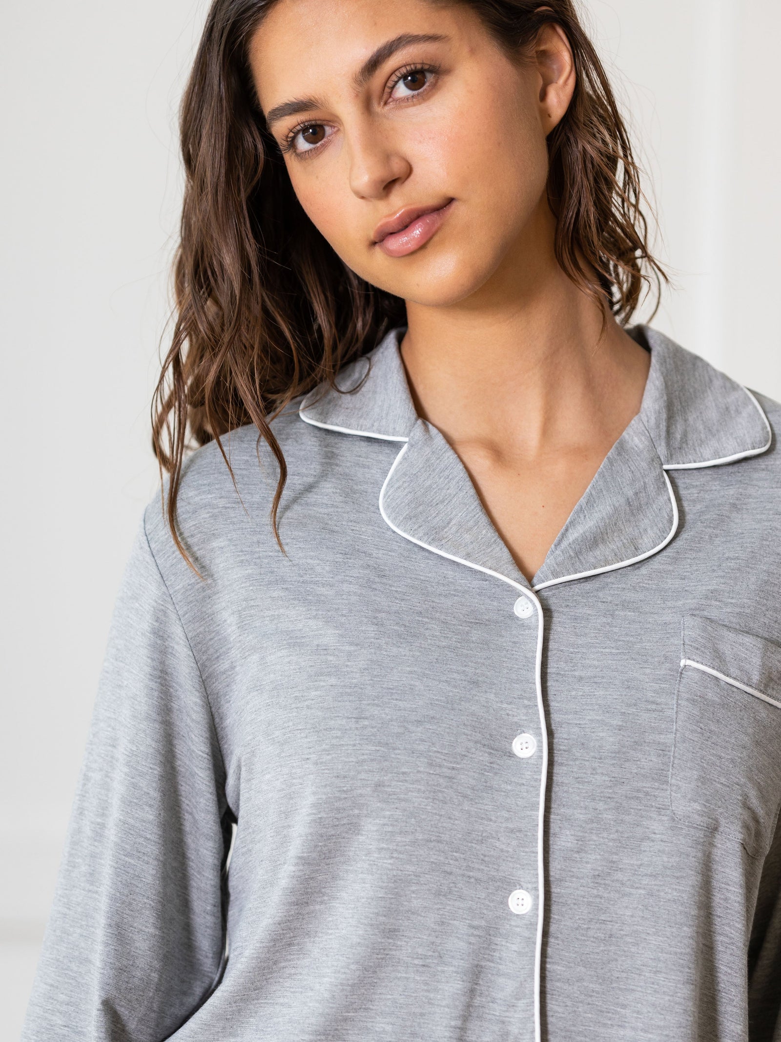A woman with wavy brown hair is wearing the Women's Bedtime Bundle by Cozy Earth, featuring a light gray pajama top with white piping and buttons. She stands in front of a plain white background, looking at the camera with a neutral expression.