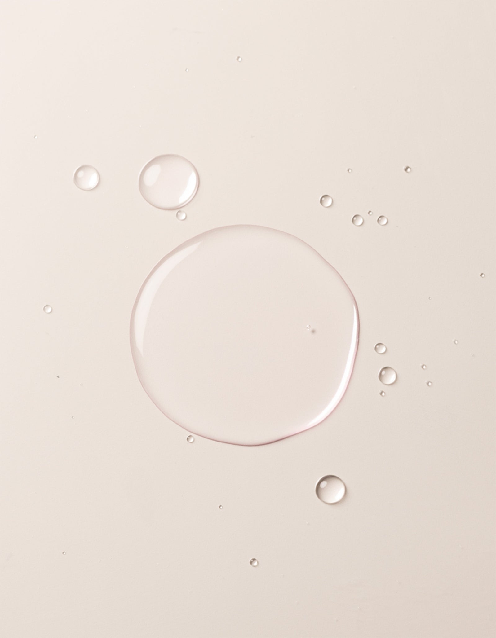 A close-up image showcases translucent liquid droplets of Cozy Earth's Balancing Face Mist on a smooth, off-white surface. The droplets vary in size, with one large central droplet surrounded by several smaller ones, creating a clean and minimalistic appearance.