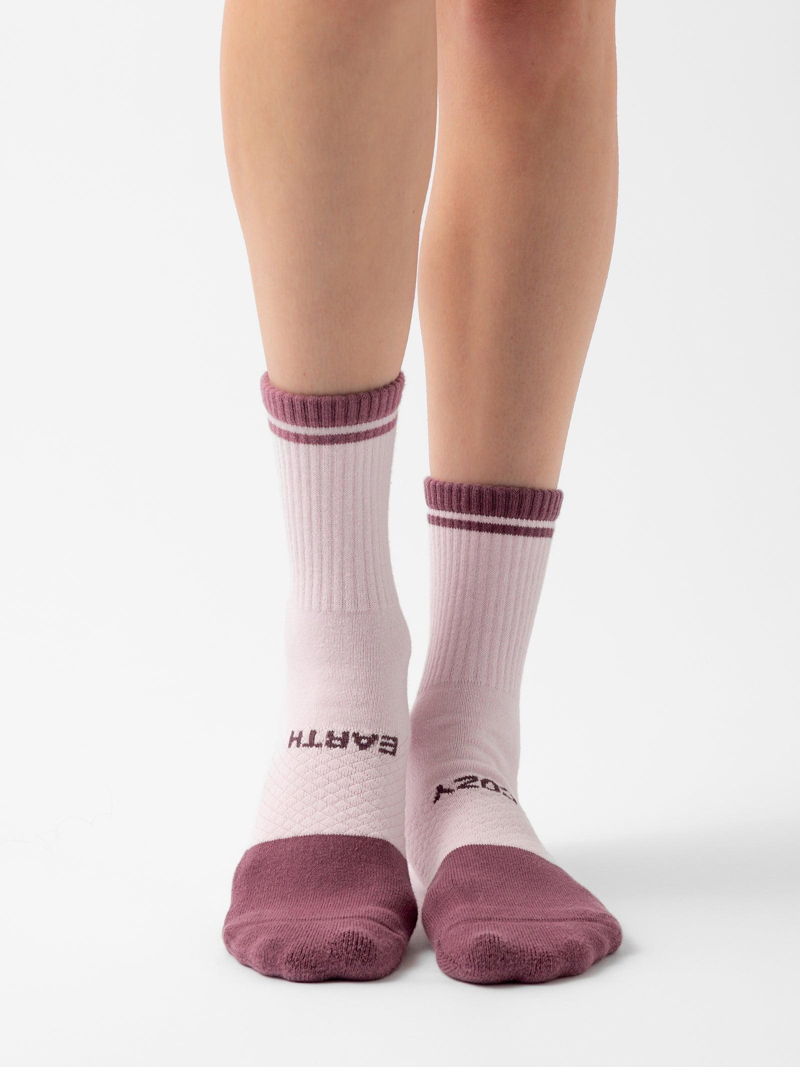 A person wearing mismatched pink socks with maroon accents from the Essential Calf Sock 4-Pack by Cozy Earth. The left sock displays the word "EARTH" on the side, while the right one features "COZY." These socks have ribbed tops and toes, and the photo is set against a plain white background.