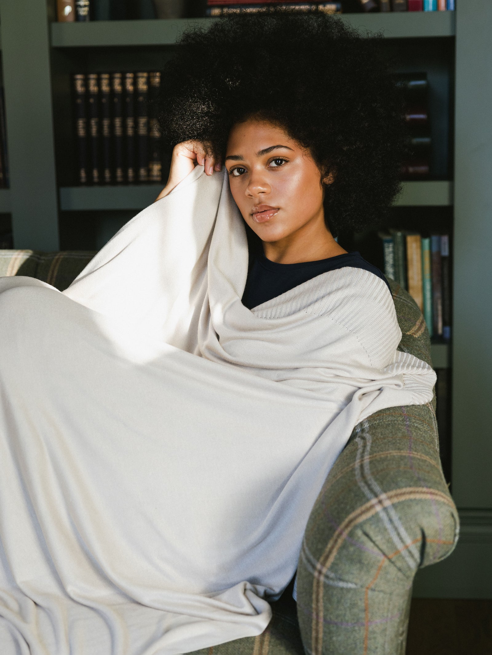 A woman is sitting reclined on a couch. She is wrapped in a Mini-Knit Blanket.