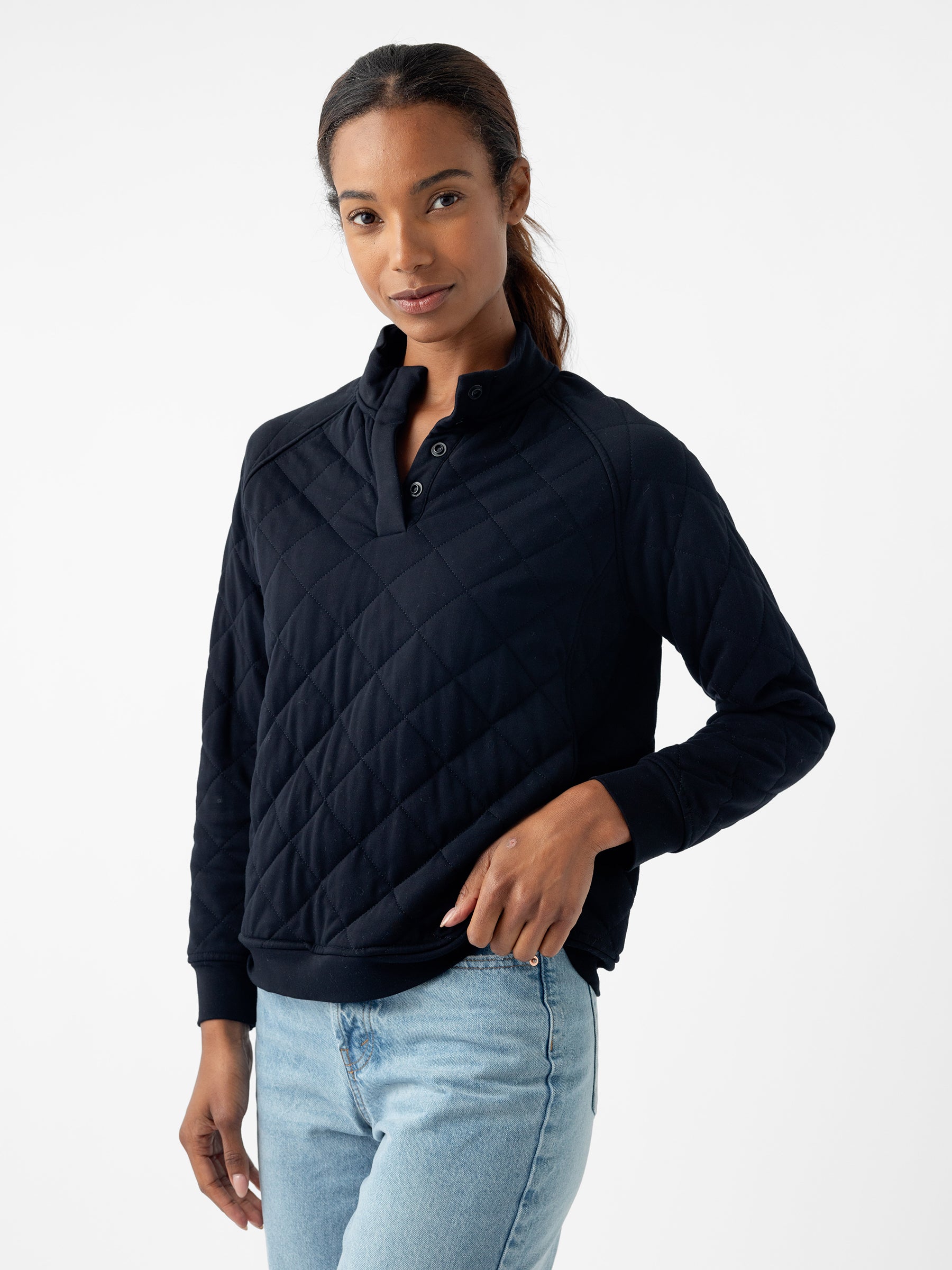 A person with long, dark hair stands against a plain white background, wearing a navy blue Women's Ultra-Soft Bamboo Quilted Snap Pullover from Cozy Earth and light blue denim jeans. They have a neutral expression and their left hand rests near their waist. |Color:Black