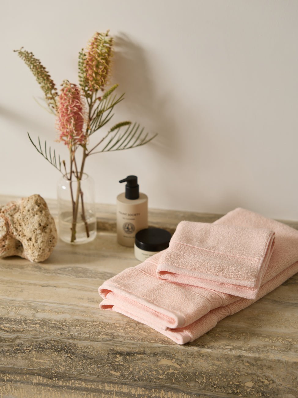 Cozy Earth's Luxe Bath Towel Set is neatly folded on a marble countertop next to a lotion bottle and a small jar. A clear vase with dried flowers enhances the decor, while a nearby textured stone adds to the natural aesthetic.