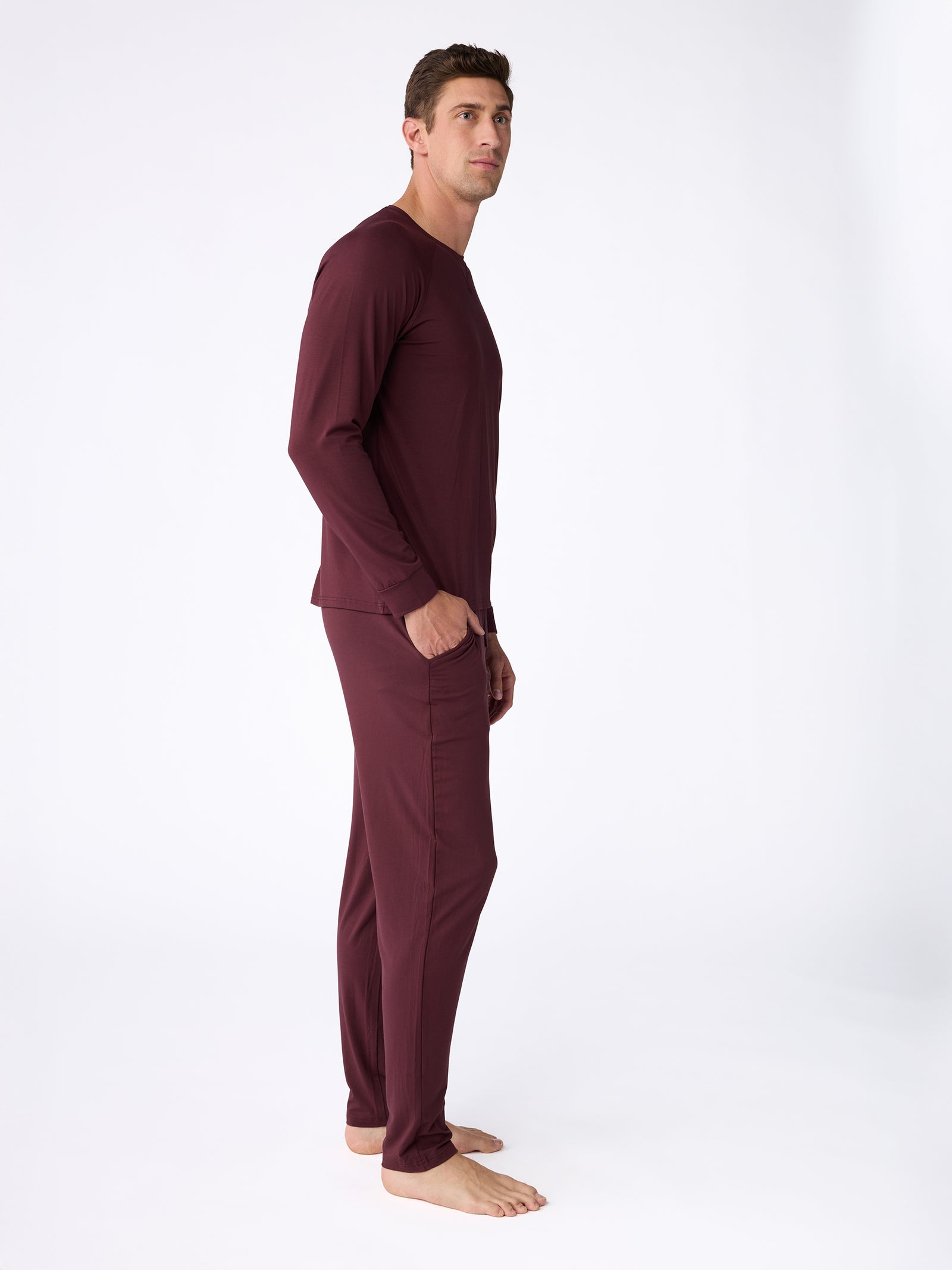 A man wearing the Cozy Earth Men's Stretch-Knit Bamboo Long Sleeve pajama set in a burgundy hue stands barefoot against a plain white background, gazing upward and to the left with his hands tucked into his pockets.
