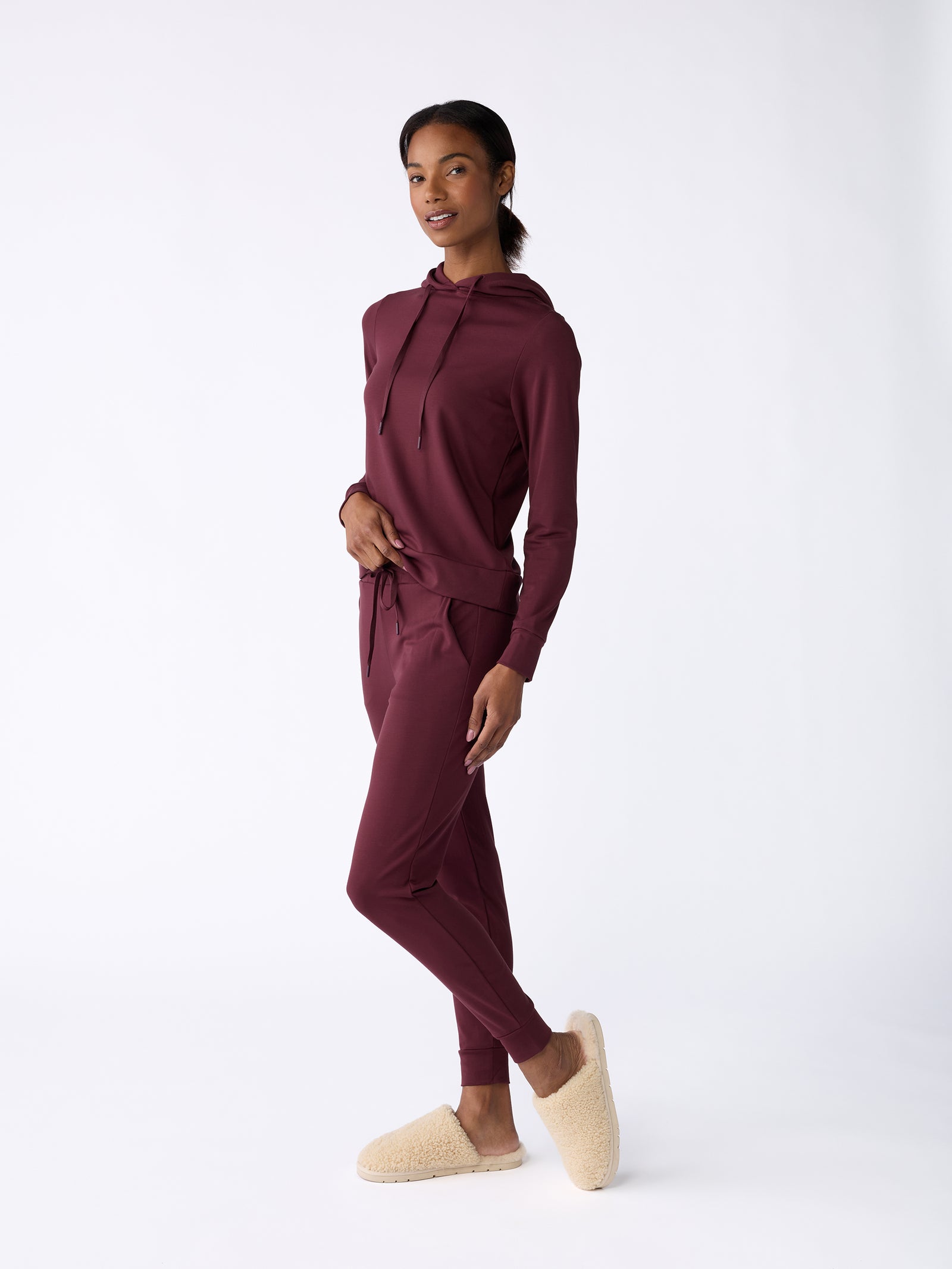A person smiling, dressed in a maroon hoodie and Cozy Earth's Women's Bamboo Jogger Pant, poses confidently against a white background. They complete the look with cozy, beige slippers.