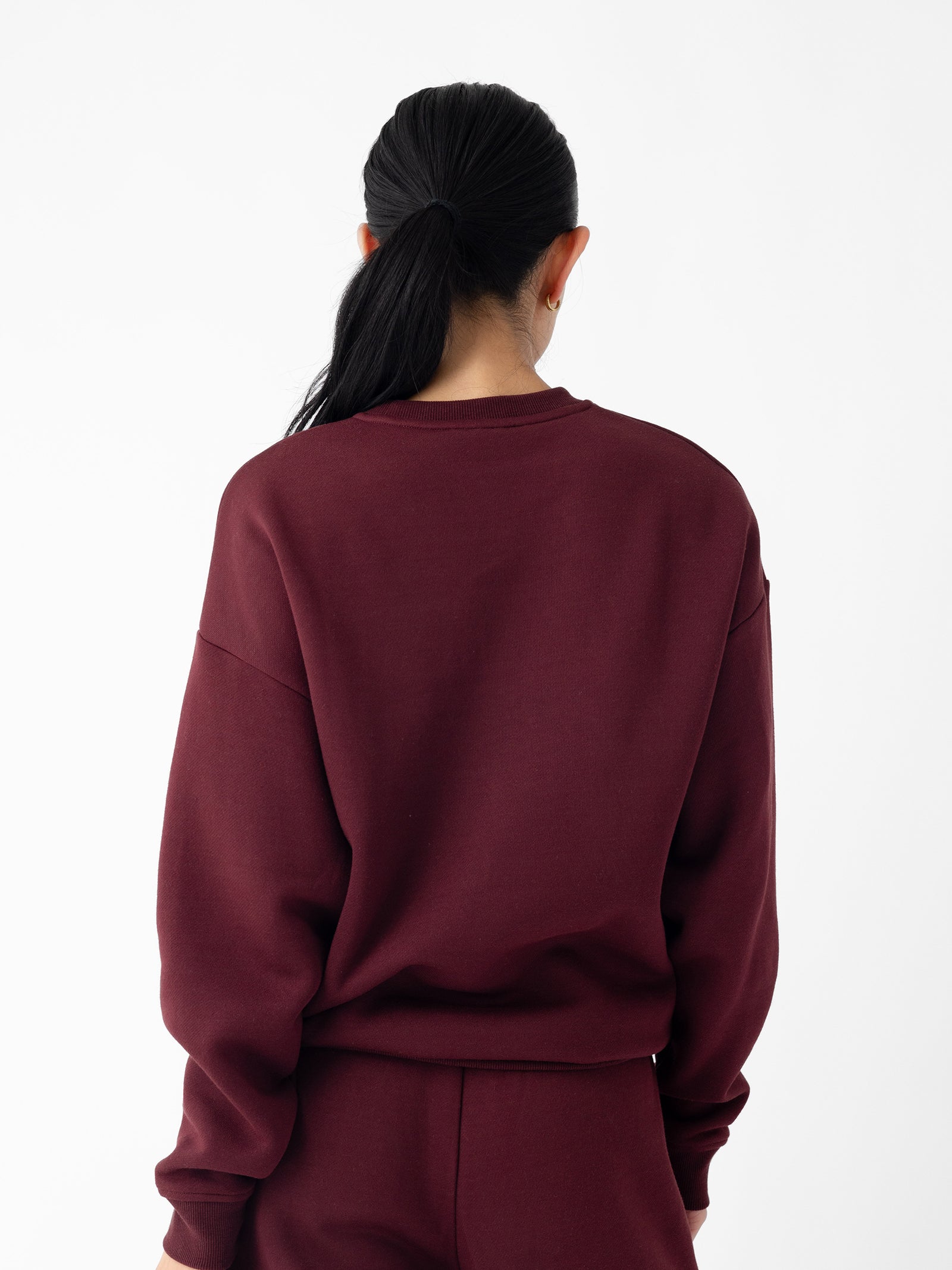 A person with long black hair is shown from the back, wearing a maroon Cozy Earth Women's CityScape Crewneck and maroon pants. The background is plain white.