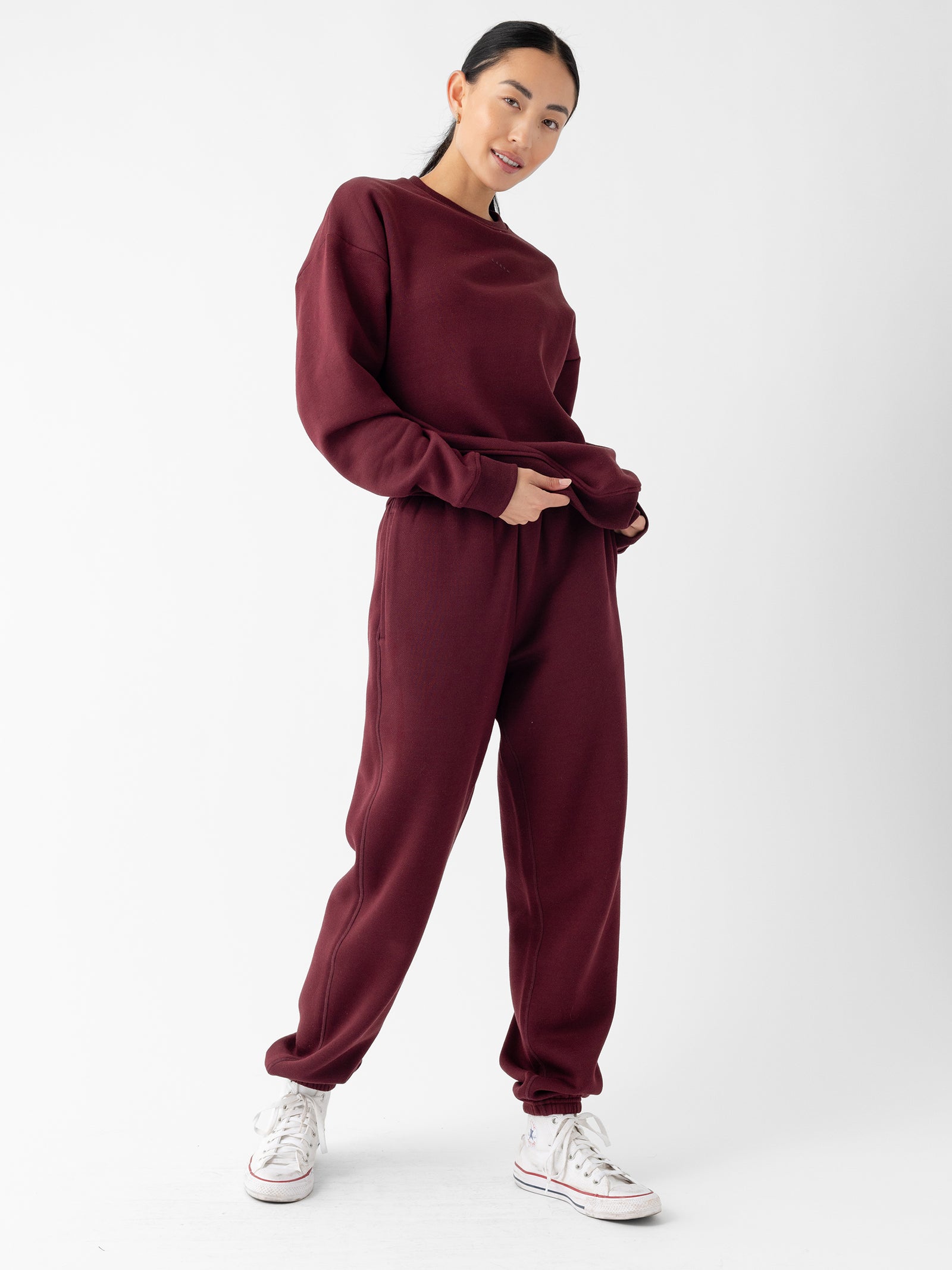A person stands against a plain white background, wearing a matching burgundy sweatshirt and Cozy Earth's Women's CityScape Sweatpant set. They have long, dark hair tied back and are smiling slightly, with their hands in the pockets of the sweatpants. They are also wearing white sneakers.