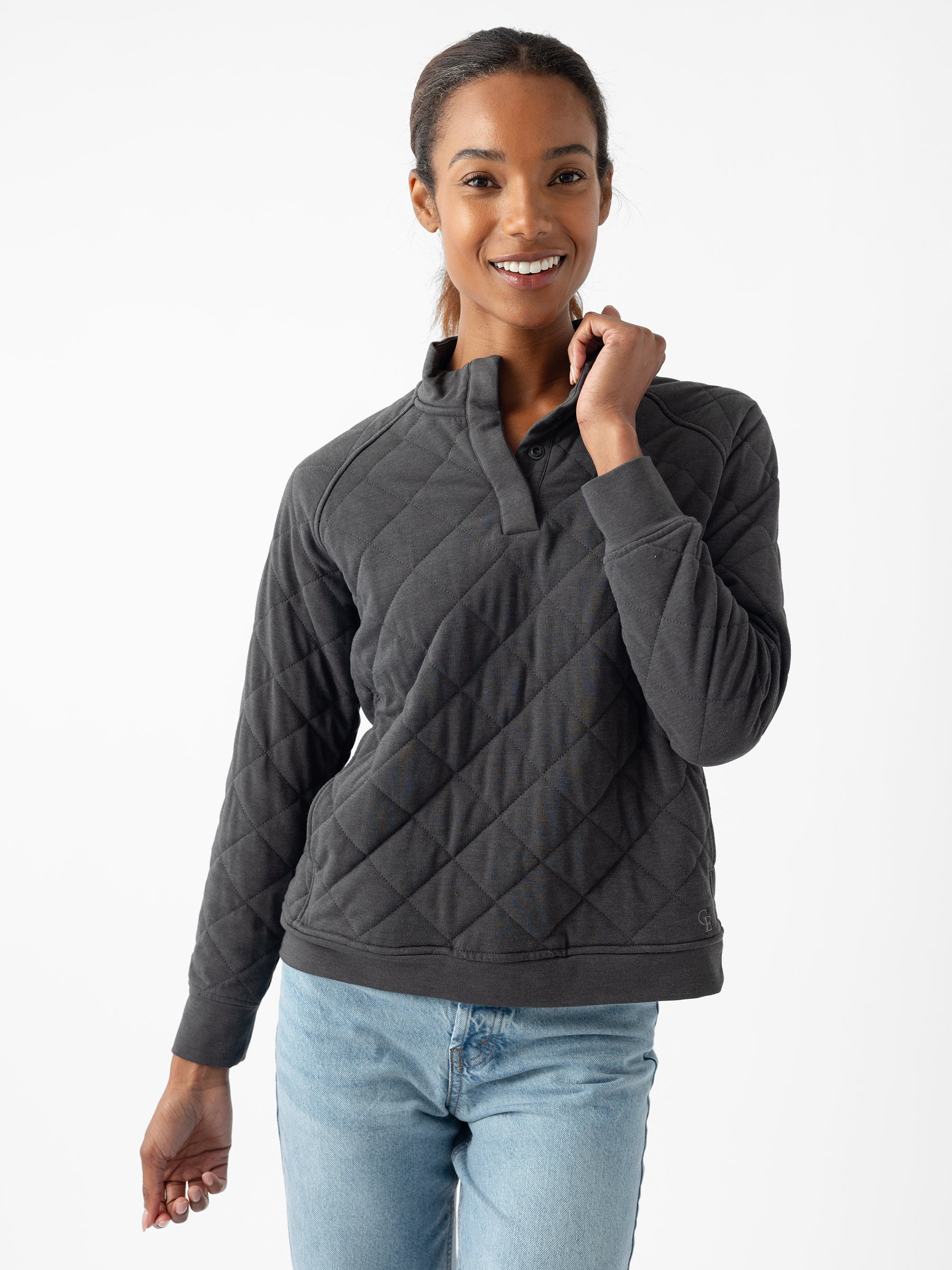 A person with dark hair is smiling at the camera, wearing a Cozy Earth Women's Ultra-Soft Bamboo Quilted Snap Pullover and blue jeans. Their right hand is raised, gently grasping the neckline of the pullover. The background is plain white. |Color:Charcoal