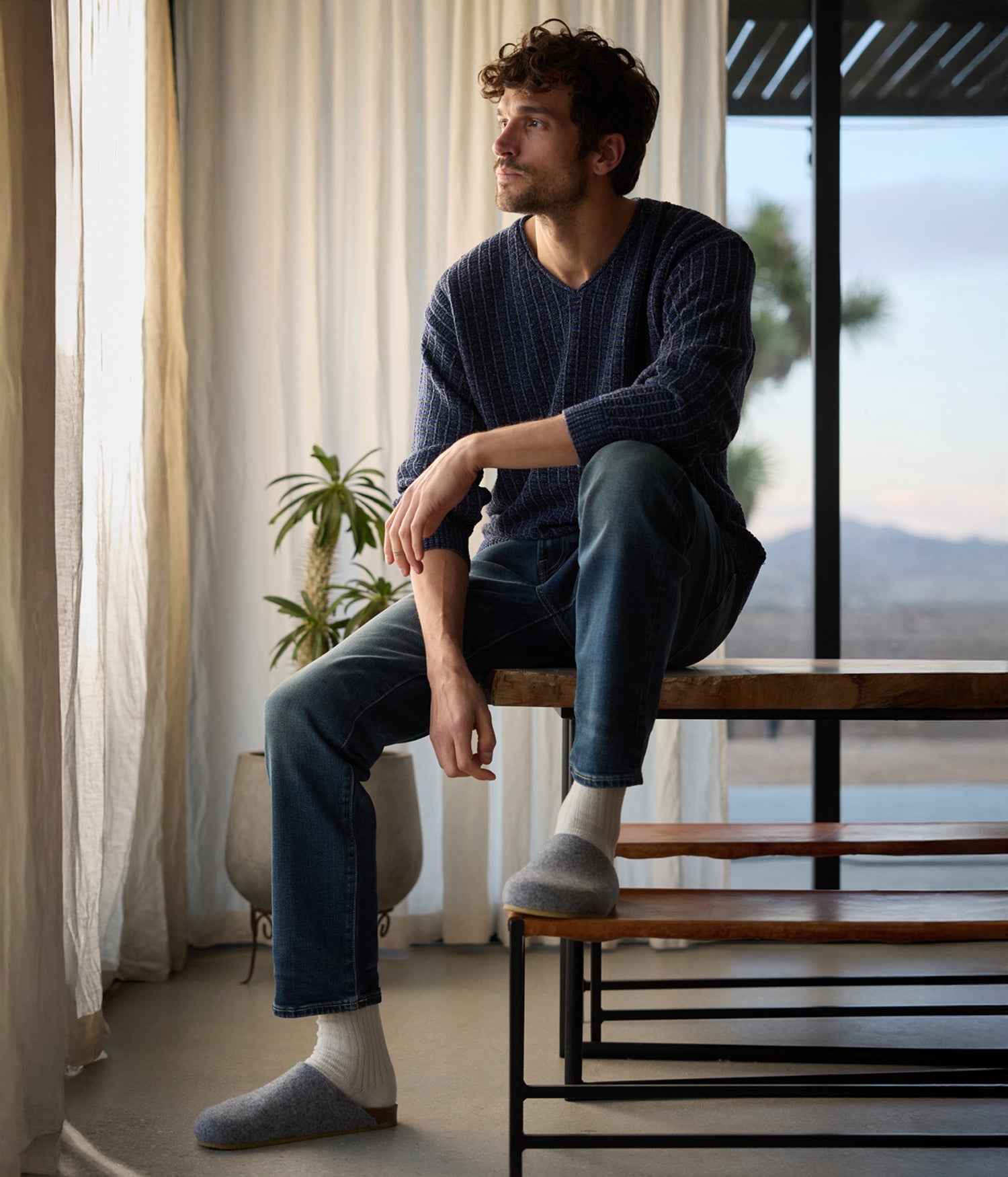 A curly-haired man sits on a wooden bench near large windows, glancing to the side. He wears a blue sweater, jeans, gray socks, and Lakehouse Clogs by Cozy Earth. Indoor plants surround him as natural light fills the room.