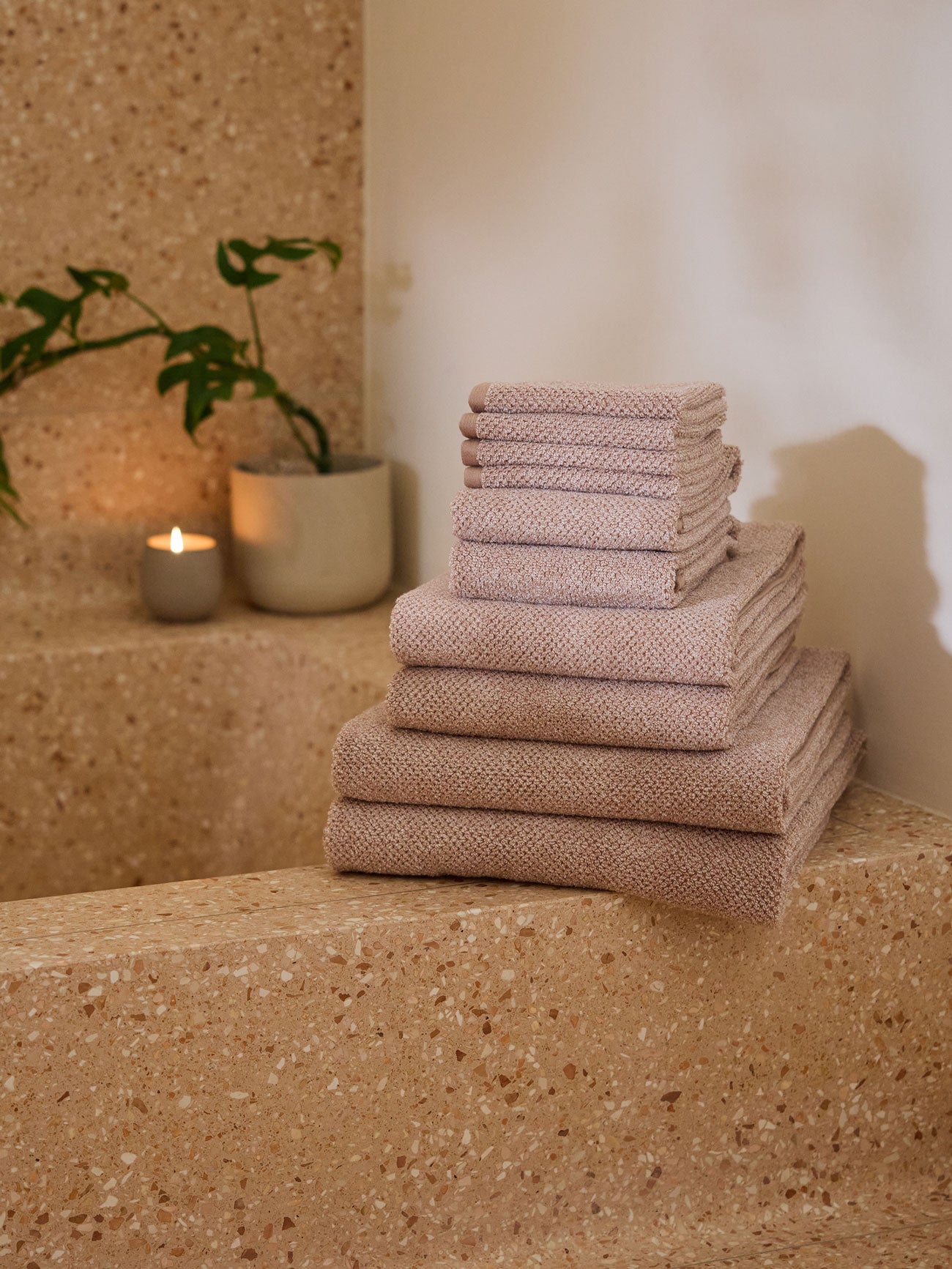 A stack of Nantucket Towels in Heathered Clay is sitting on the edge of a stone bathtub. A small plant and lit candle are sitting on the opposite edge.