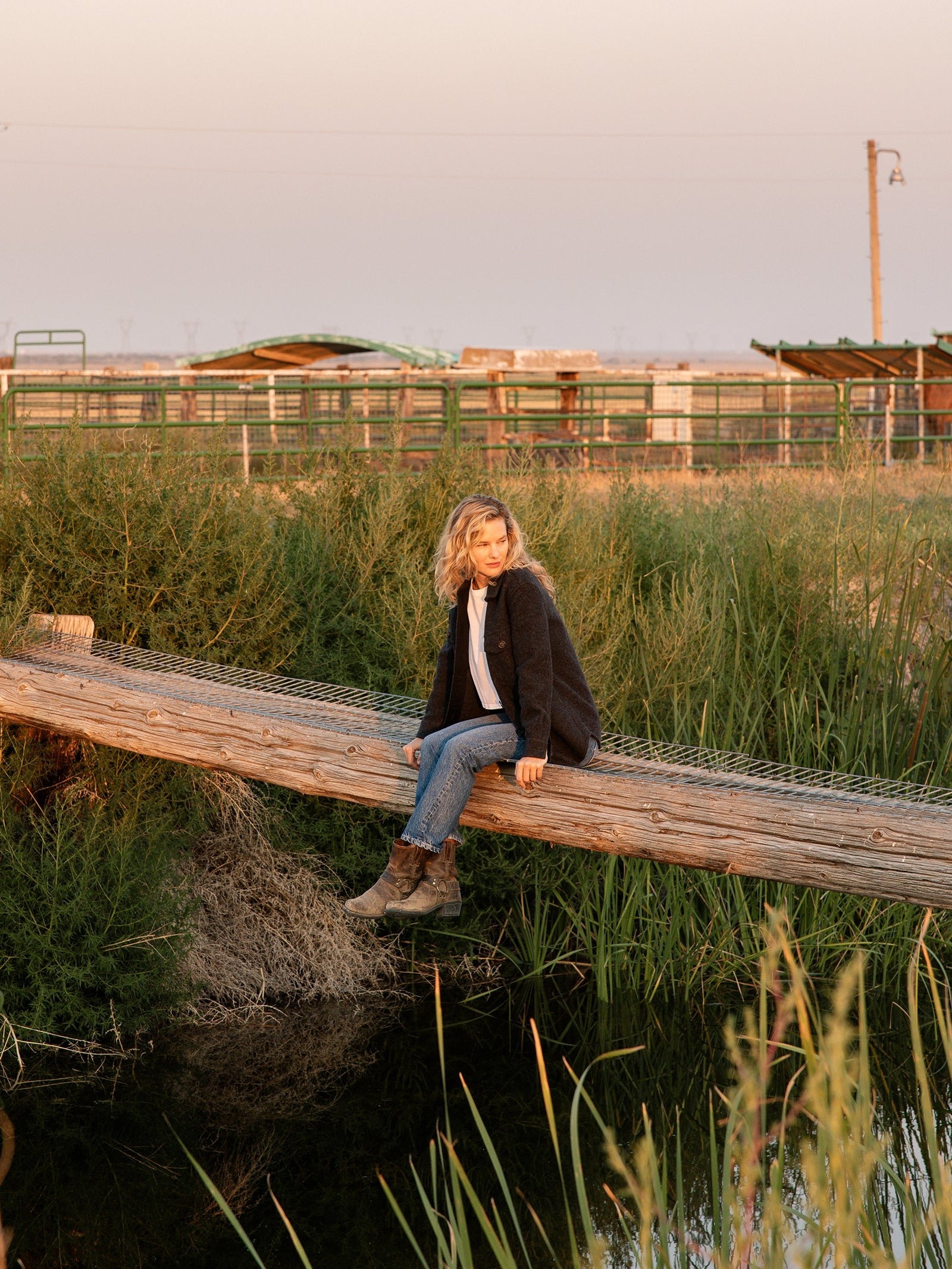 A person with short, wavy blonde hair sits on a wooden beam over a grassy and watery landscape. They wear a Cozy Earth Women's Boucle Shacket, a white shirt, blue jeans, and boots. In the background, there are green and brown agricultural structures under an evening sky.