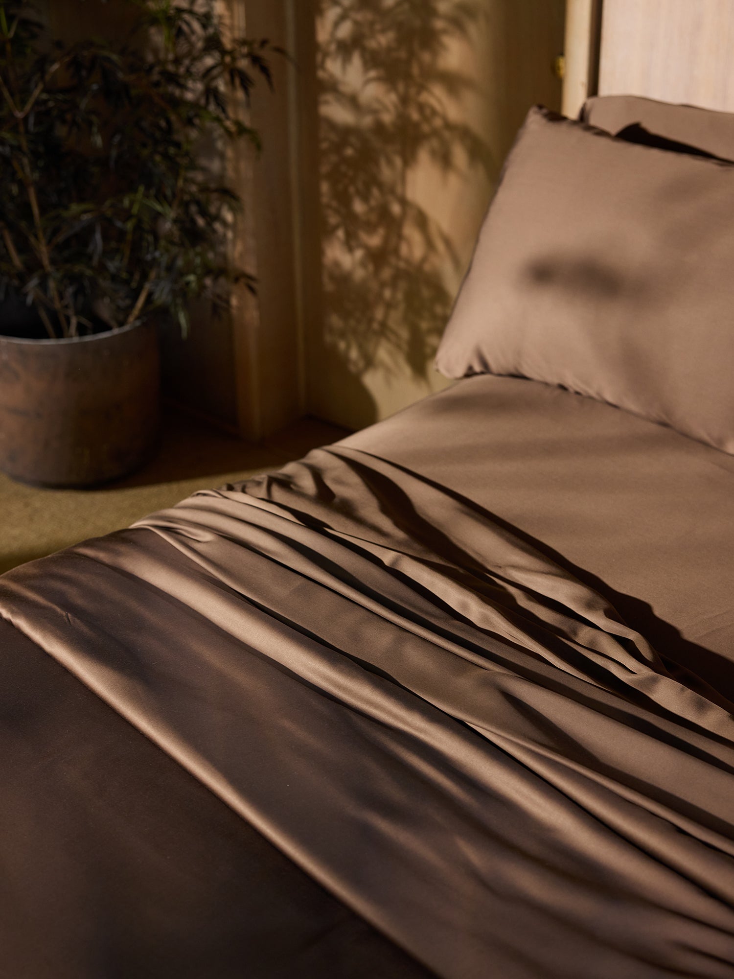 A neatly made bed with Cozy Earth's Bamboo Flat Sheets in a warm brown hue and matching pillows is bathed in sunlight. To the left, a potted plant adds a touch of greenery. Shadows of the plant's leaves are cast on the wall and bedding, creating a cozy and inviting atmosphere.