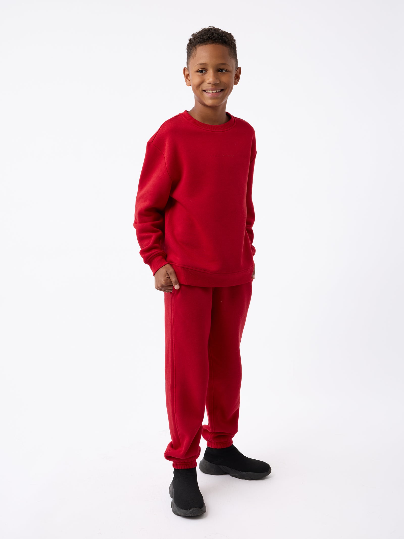 A young boy smiles while dressed in the Kid's CityScape Sweatpant, a matching red sweatshirt and sweatpants set from Cozy Earth, paired with black shoes. He stands against a plain white background.