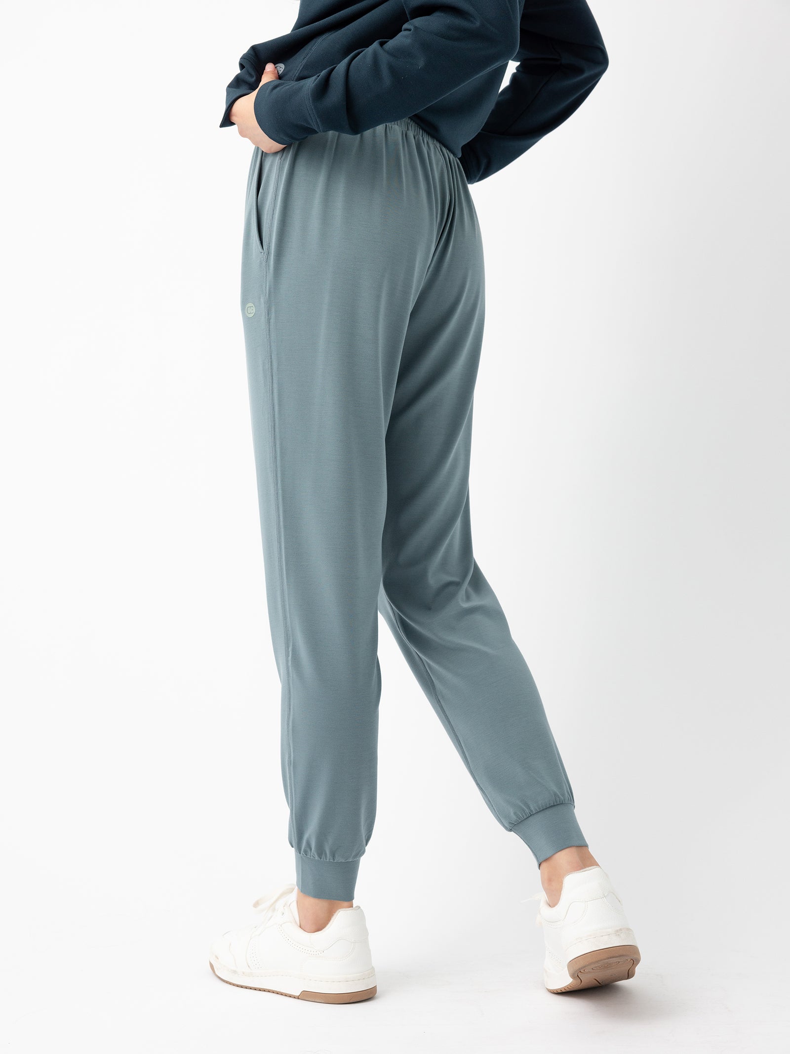 A person stands with one hand in their pocket, wearing Women's Studio Jogger pants by Cozy Earth in a teal color, a dark long-sleeve top, and white sneakers. The joggers feature a relaxed fit with an elastic waistband and cuffs at the ankles. The person is posing against a plain white background.