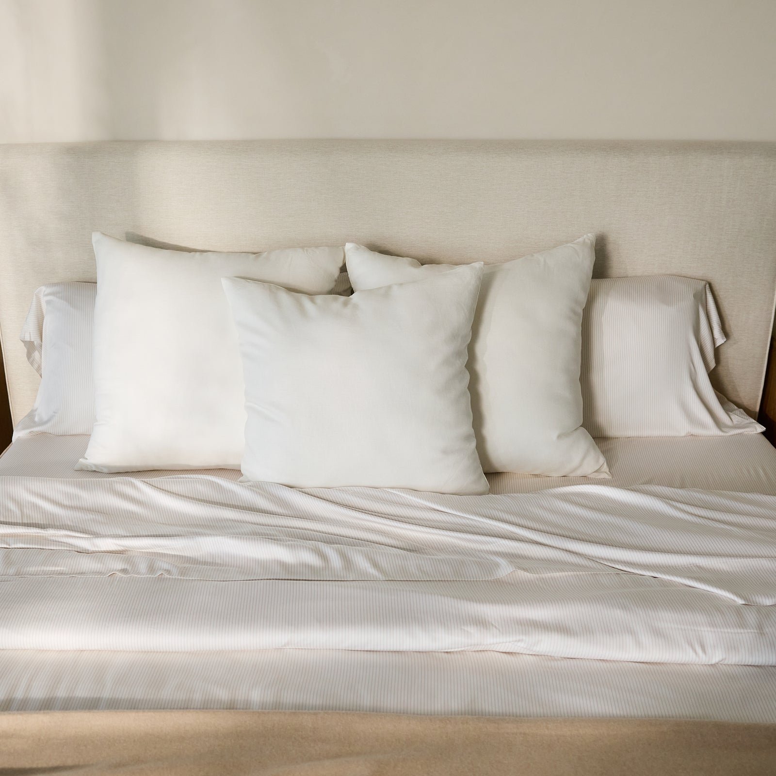 A neatly made bed, featuring a beige fabric headboard, is adorned with four white pillows and topped with the Bamboo Duvet Cover by Cozy Earth. Sunlight softly illuminates the bedding, creating a serene and inviting atmosphere.