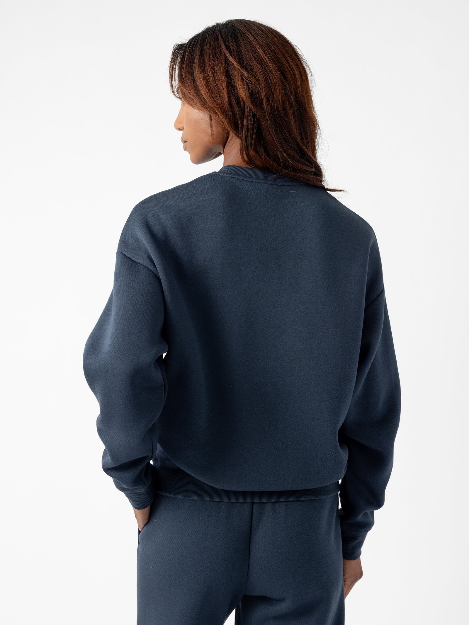 A person with shoulder-length, dark brown hair is seen from the back, wearing a dark blue Women's CityScape Crewneck by Cozy Earth and matching pants. They are standing against a plain white background.