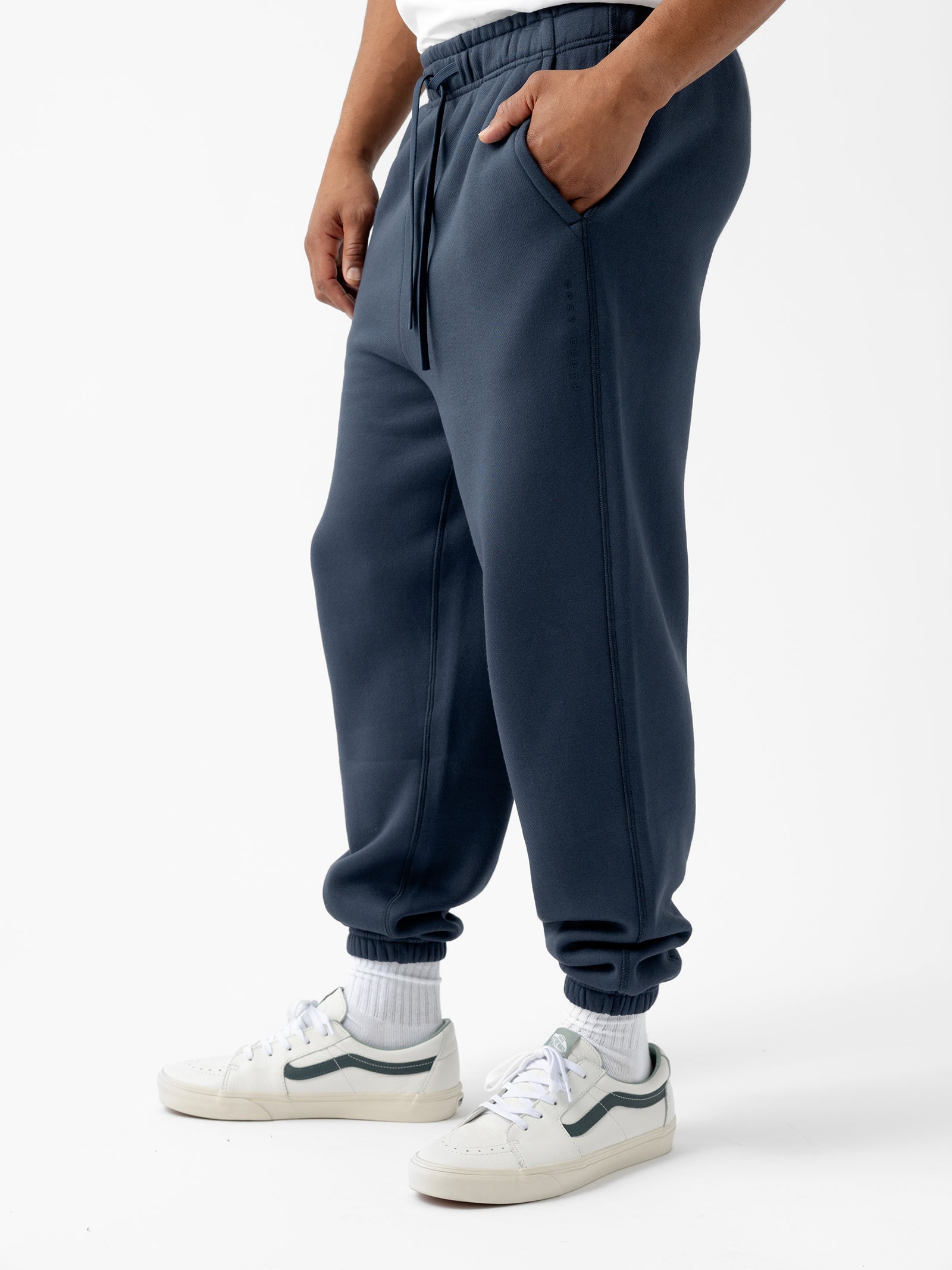 A person wearing Cozy Earth's Men's CityScape Sweatpant in dark blue with pockets, a white T-shirt, white socks, and white sneakers with dark accents stands against a plain white background. The person's hands are in the pockets of the sweatpants.