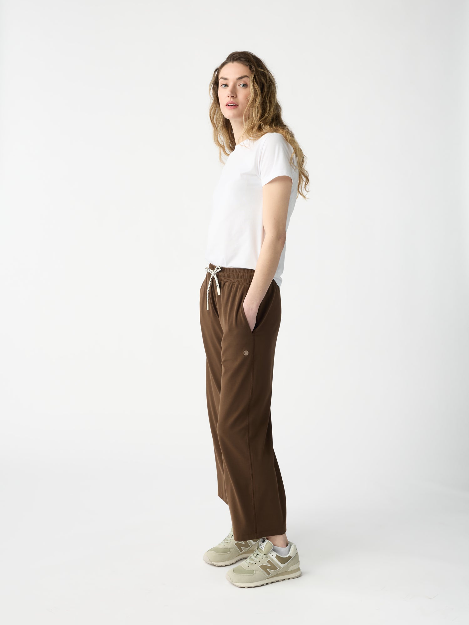 A person with long, wavy hair stands in profile against a white backdrop, wearing a white T-shirt, Cozy Earth's Women's Studio Cropped Wide Leg Pant in brown with a drawstring, and beige sneakers. Their hands are in pockets, and they have a neutral expression.