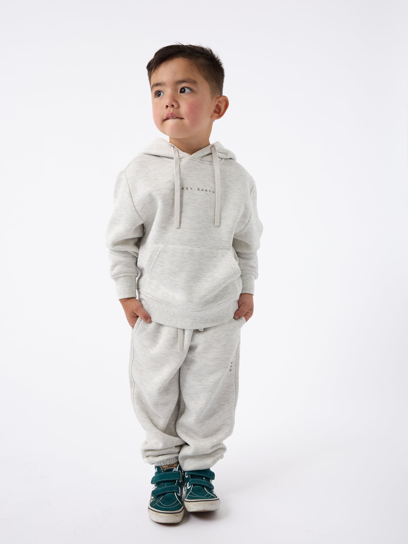 A young boy dressed in Cozy Earth's Kid's CityScape Sweatpant and teal sneakers stands against a plain white background, looking slightly to his left with his hands tucked into the pockets of his hoodie.