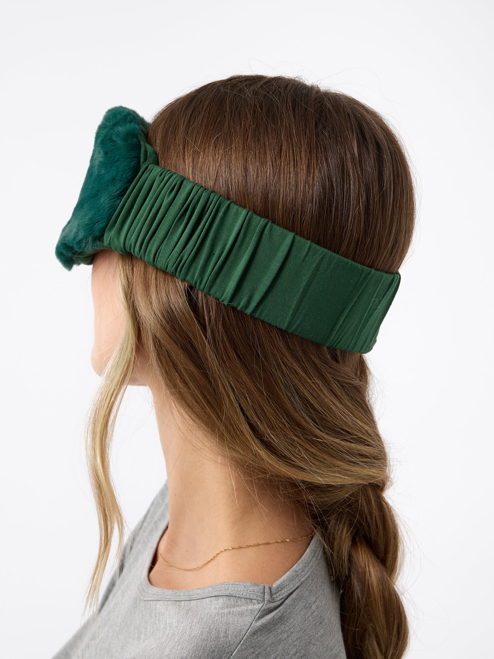 A woman wears the Cozy Earth Faux Fur Sleep Mask in Jade. Her head is turned to show the elastic band that wraps around her head.