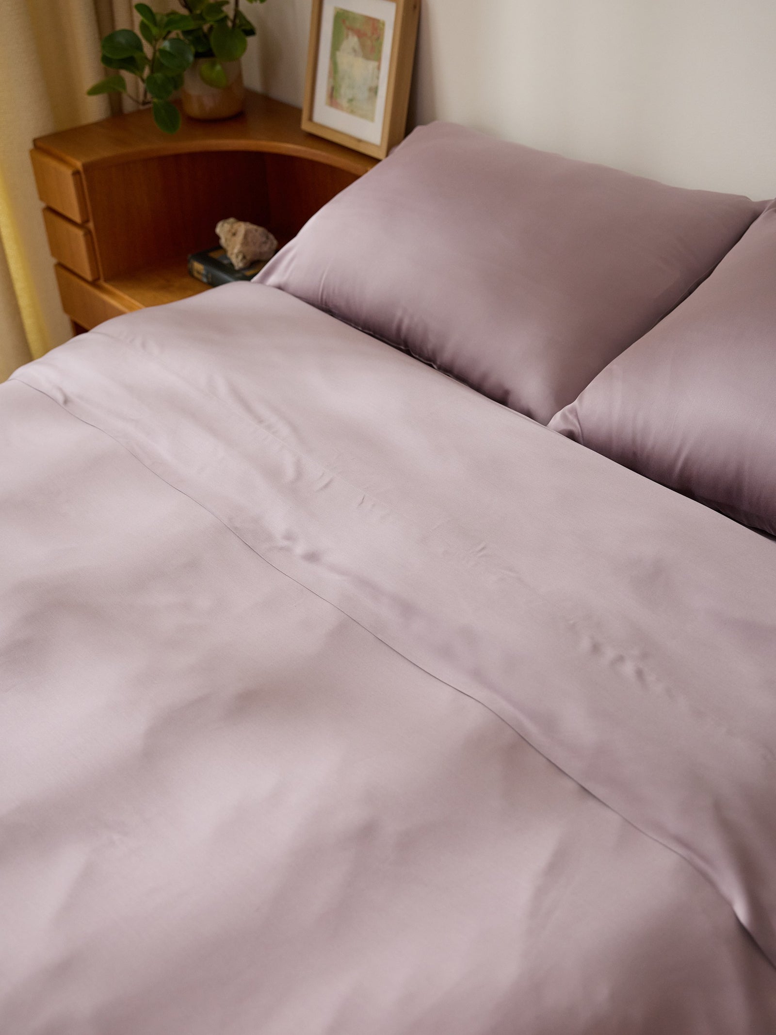 A neatly made bed with Cozy Earth's Bamboo Flat Sheet in lavender and two matching pillows. A wooden nightstand beside the bed holds a small plant, a framed picture, and other decor items against a white wall.