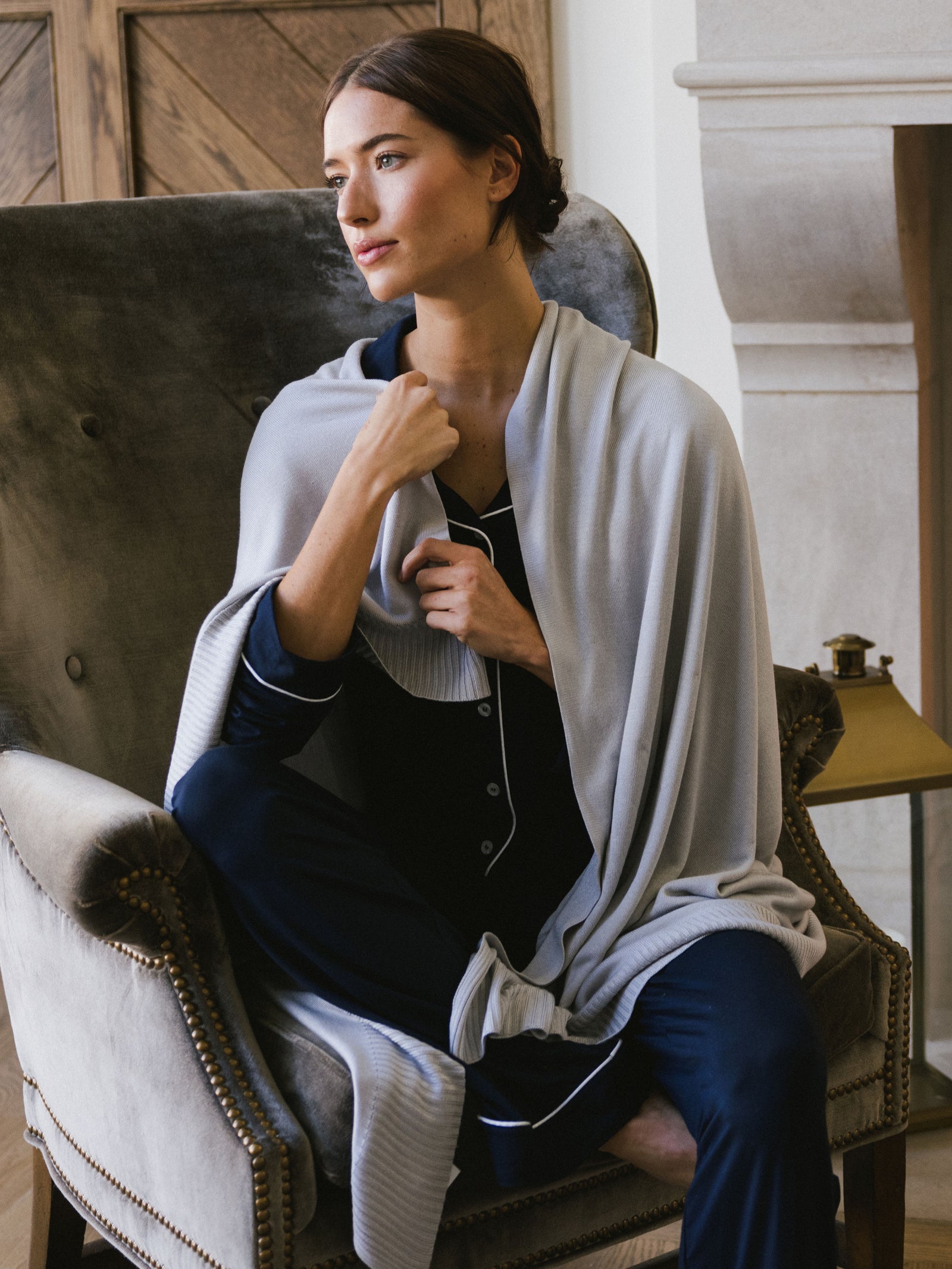 A woman is sitting on a couch. As she longingly looks out what is assumed to be a window, she warms herself with a Light Grey Mini-Knit Blanket.