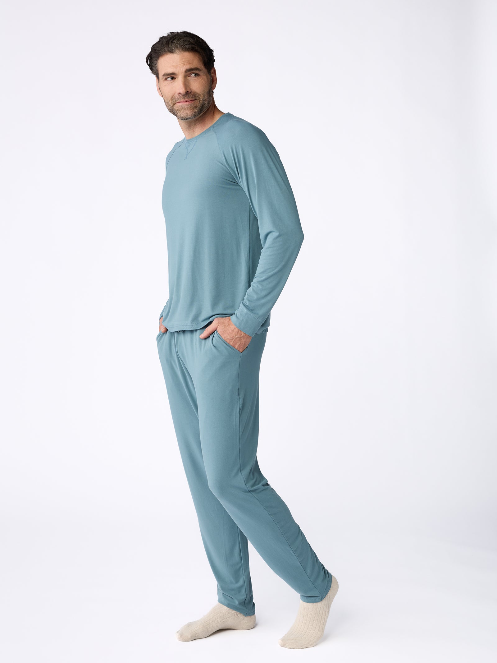 A person stands in a relaxed pose, wearing a Men's Stretch-Knit Bamboo Long Sleeve from Cozy Earth, paired with matching blue pajama pants and beige socks. They have their hands in the pockets of the pajama pants against a plain white background.