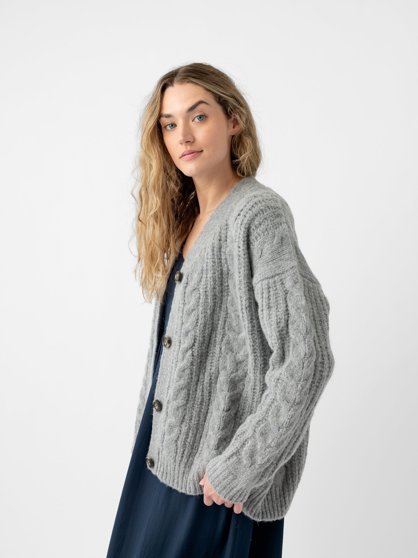 A person with long, curly hair is wearing the Oversized Cable Knit Cardigan by Cozy Earth in gray over a dark dress. They are standing against a plain, white background and looking towards the camera.