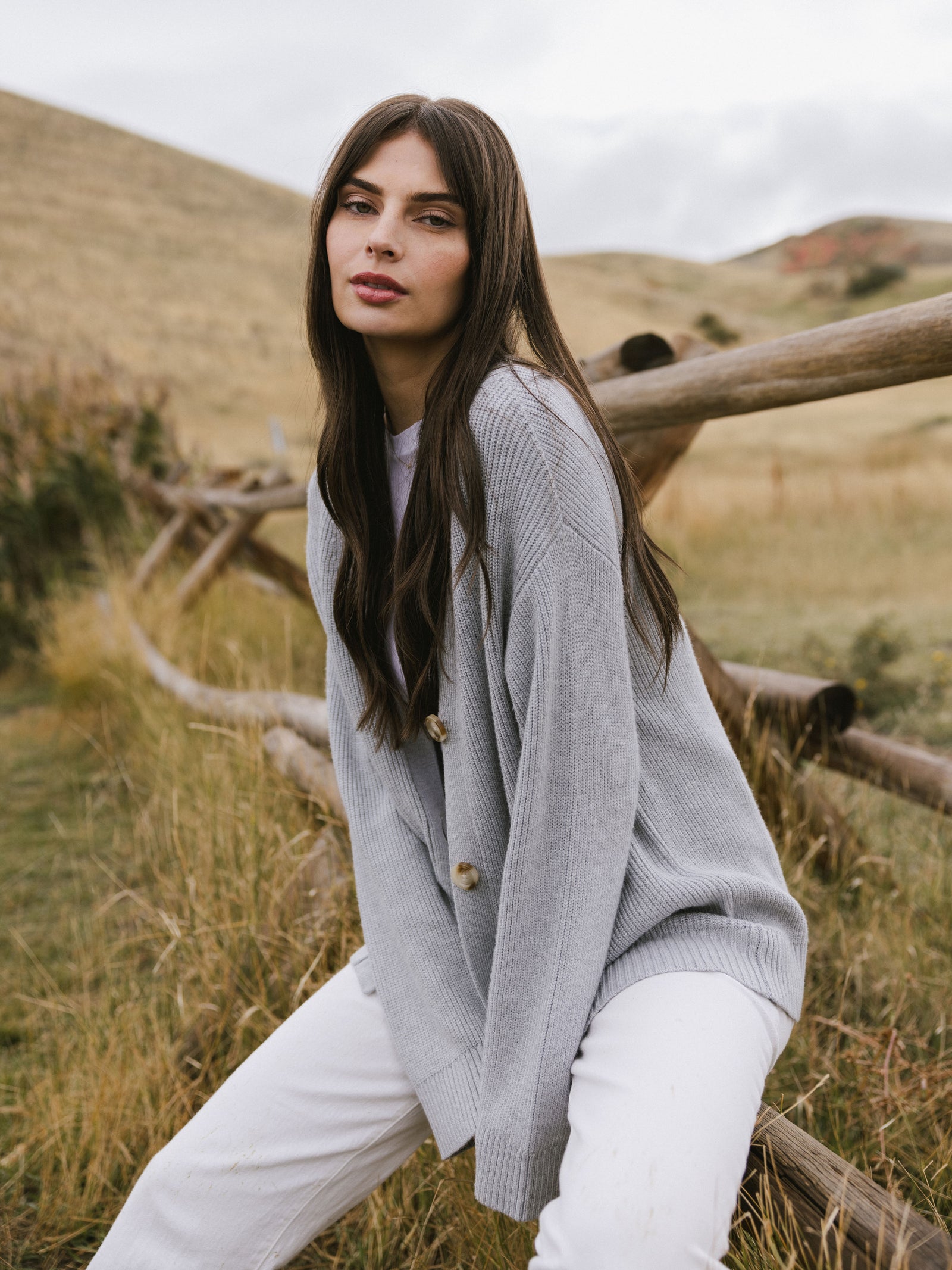 A woman wearing Cozy Earth Classic Cardigan in grey sits on a fence in a field