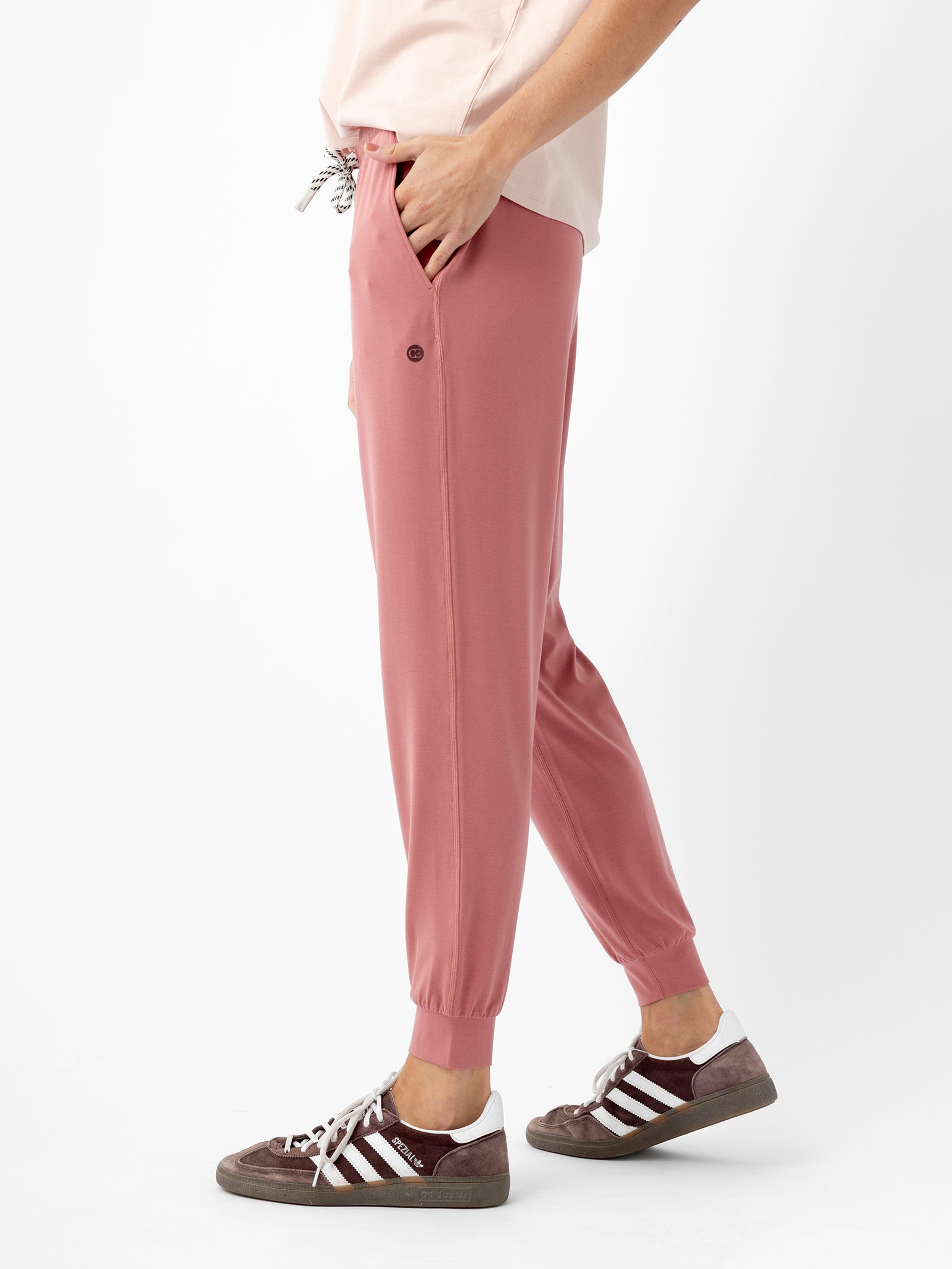 A person dressed in a light pink top and Cozy Earth's Women's Studio Jogger pants in pink is seen from the side. The joggers feature a drawstring and pockets, and the individual is wearing gray and white sneakers. The background is white.