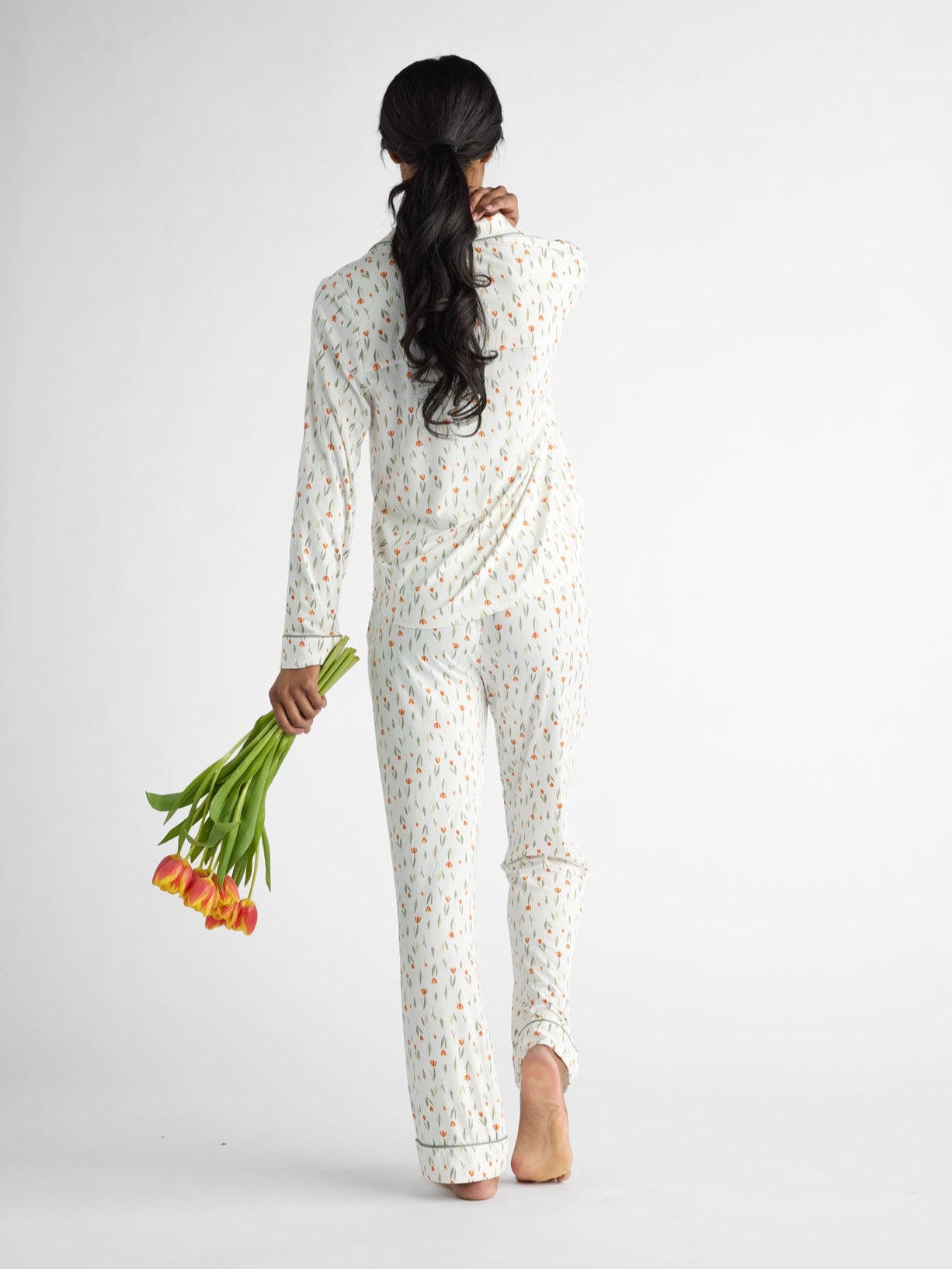 Wearing a Women's Bamboo Stretch-Knit Long Sleeve Pajama Top by Cozy Earth, a person with long hair walks barefoot away from the camera, holding orange tulips against a plain white background.