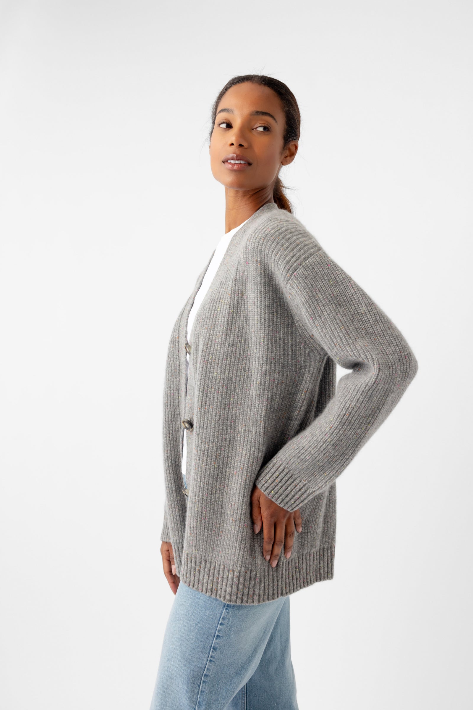 A person wearing an oversized Sunday Cashmere Cardigan from Cozy Earth in gray over a white shirt and light blue jeans stands against a plain white background, looking to the side with one hand on their hip.