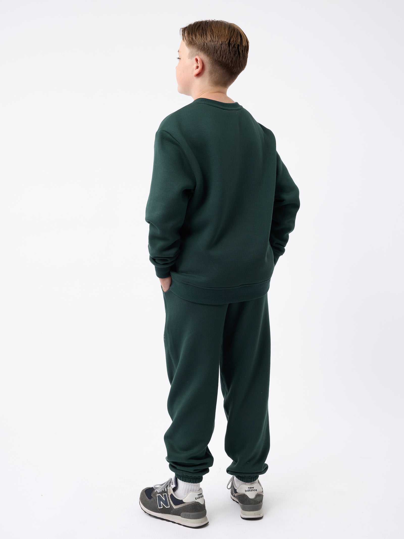 A boy dressed in a Cozy Earth Kid's CityScape Sweatpant, standing with his back to the camera and looking to the side. He is wearing gray sneakers against a plain white background.