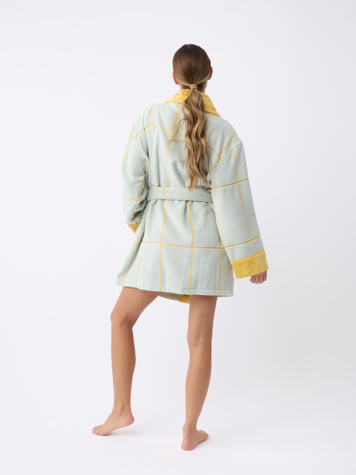 A barefoot person with long hair stands facing away, wearing a Cozy Earth Windowpane Resort Robe in light green plaid with yellow accents, cinched at the waist. The background is plain white.