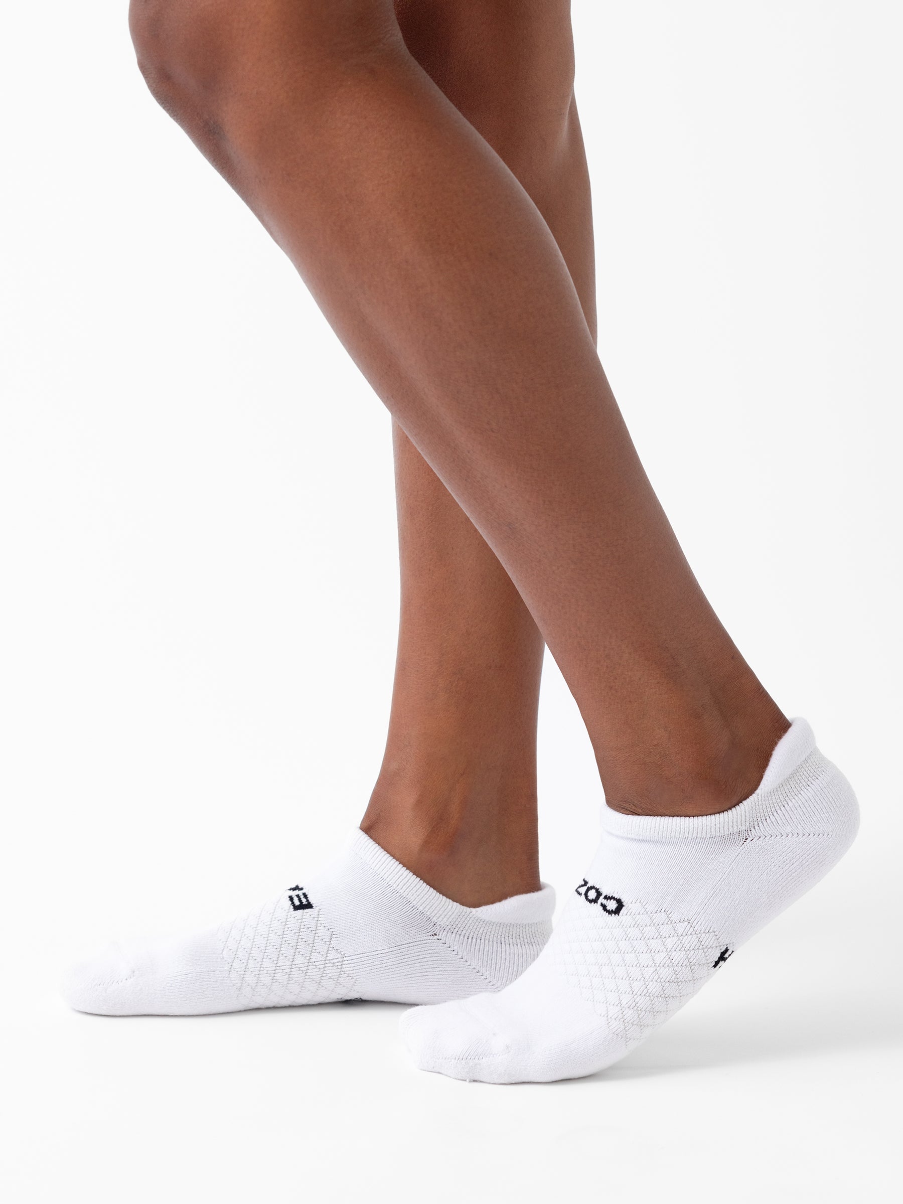 Close-up of a person's legs and feet wearing white Essential Ankle Socks from the Cozy Earth 2-Pack, featuring black text that reads "COZY EARTH" on the side. The plain white background accentuates the legs and socks. |Color:White|Count:2-Pack