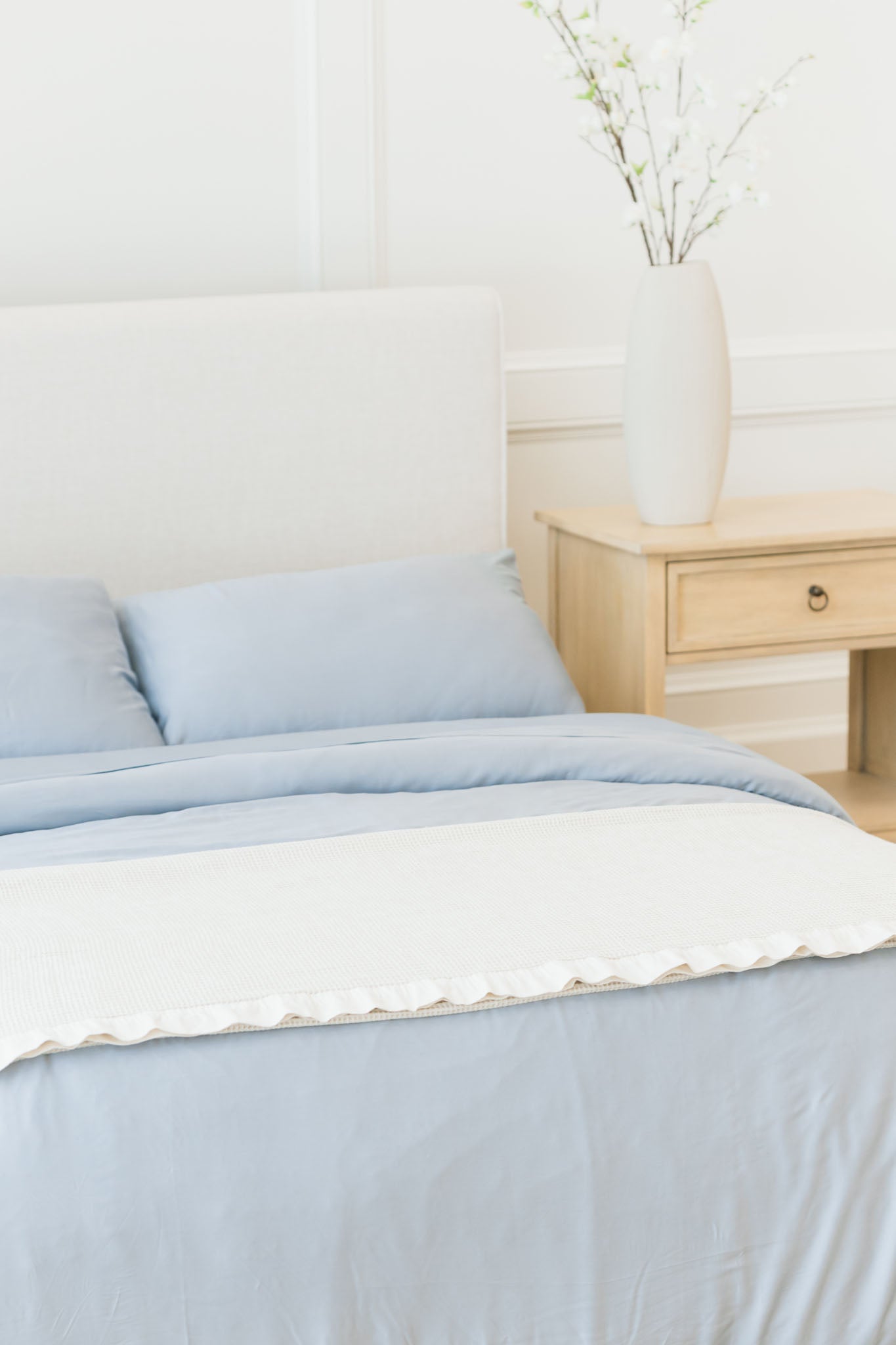 A neatly made bed with blue bedding and a Cozy Earth Waffle Blanket in white is in front of a beige upholstered headboard. Next to the bed is a wooden nightstand with a large white vase holding branches with small white flowers. The room has a light and airy feel.