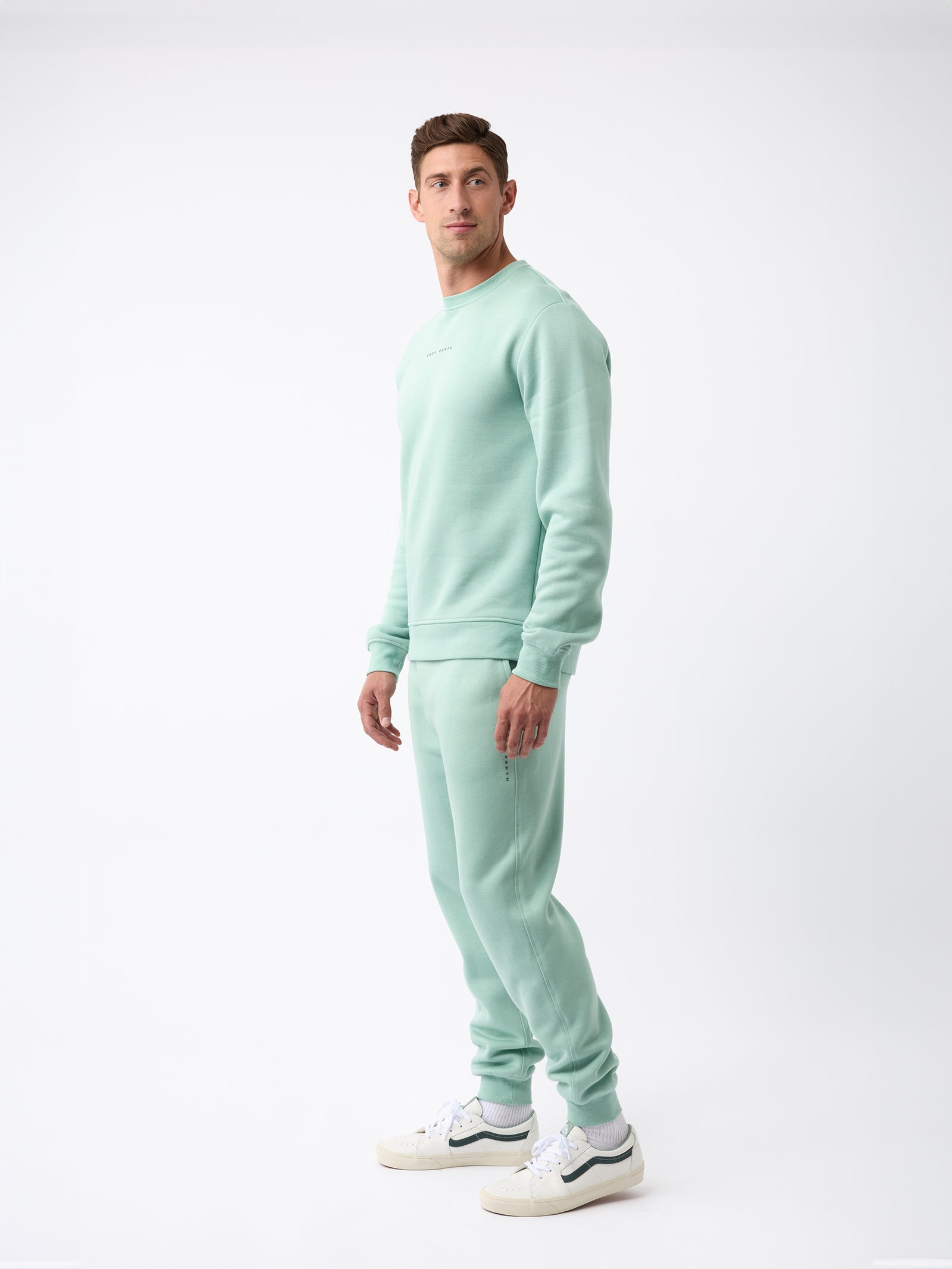 A person wearing the Men's CityScape Crewneck in a light green shade along with matching joggers stands against a plain white background. They are also wearing white sneakers from Cozy Earth.