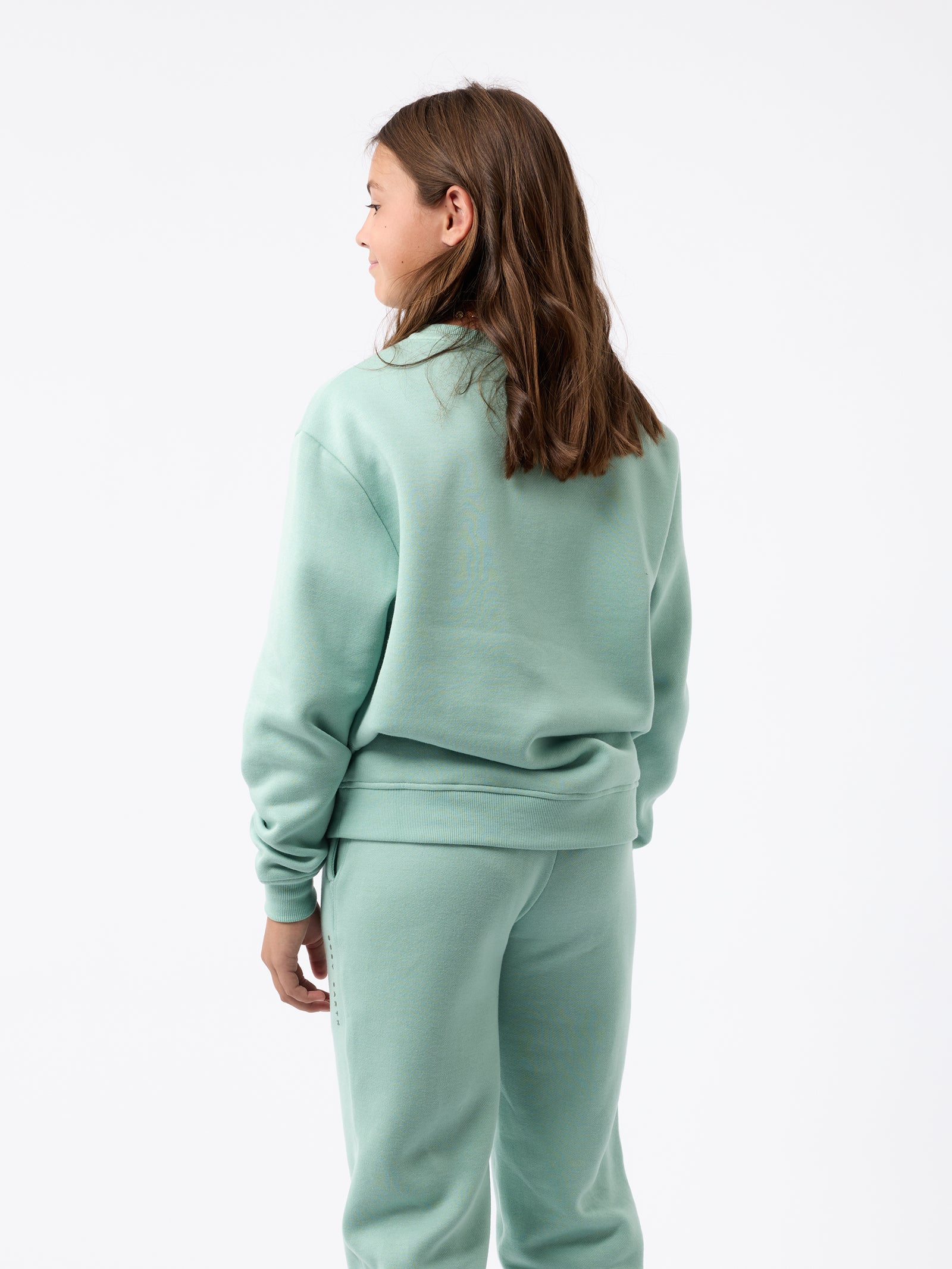A person with long brown hair is standing and facing away from the camera, wearing a light green Kid's CityScape Crewneck by Cozy Earth, along with matching sweatpants against a plain white background.